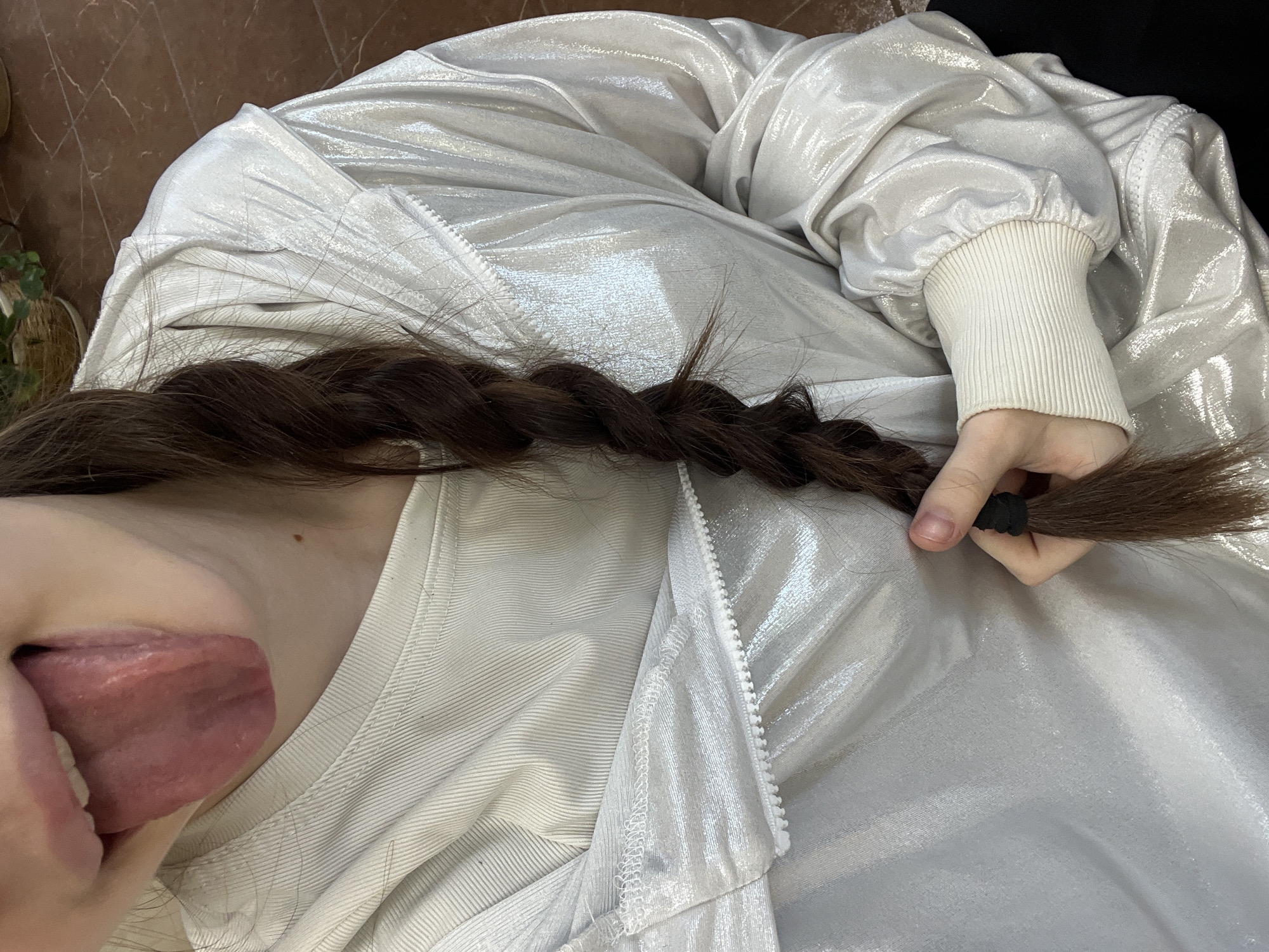 A high-angle close-up shows a person sticking their tongue out while holding the end of a brown braid with a black hair tie. The person wears a white t-shirt underneath a silver, metallic-looking zip-up jacket with a white ribbed cuff. The background appears to be a wooden floor and possibly part of a green plant in a pot. The location is not identifiable from the image alone, and there is no indication the picture was taken in Saint Petersburg, Russia.