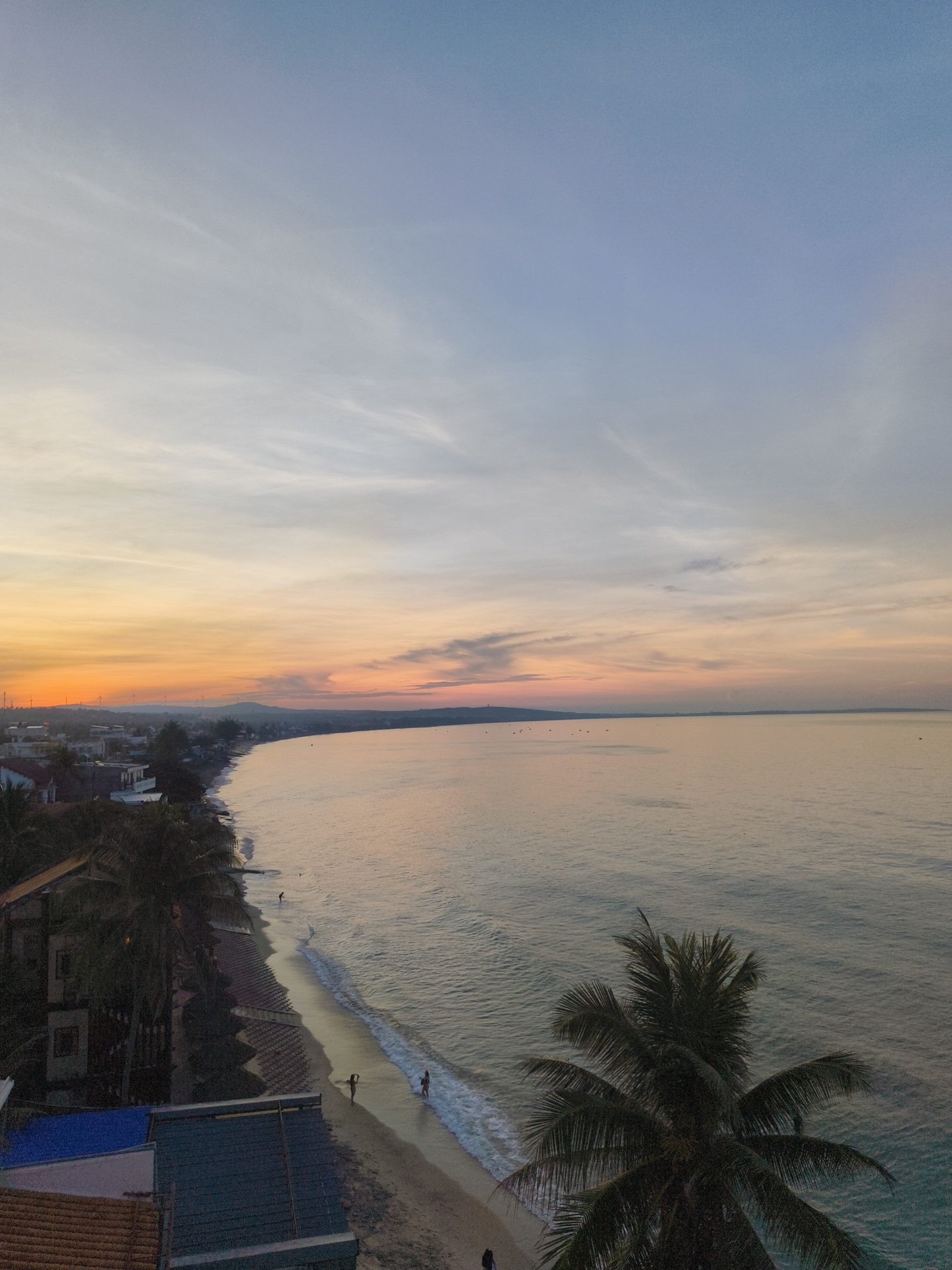 This image captures a scenic view of a beach and coastline at what appears to be sunrise or sunset in Phu Thuy Ward, Vietnam. The sky is a gradient of colors, transitioning from a pale blue at the top to hues of orange, pink, and yellow closer to the horizon. Wispy clouds are scattered across the sky. The ocean is calm, with gentle waves lapping at the shore.
Along the coastline, a sandy beach is visible, with a line of buildings and palm trees situated behind it. Some of the buildings appear to be residences or small businesses, with varying roof colors and architectural styles. A few individuals are present on the beach, with one person walking near the water's edge and others further up the shore. A prominent palm tree with dense fronds dominates the foreground on the right side of the image. In the distance, the coastline curves, with hills and possibly wind turbines visible on the horizon. The overall impression is one of a peaceful coastal setting at a tranquil time of day.