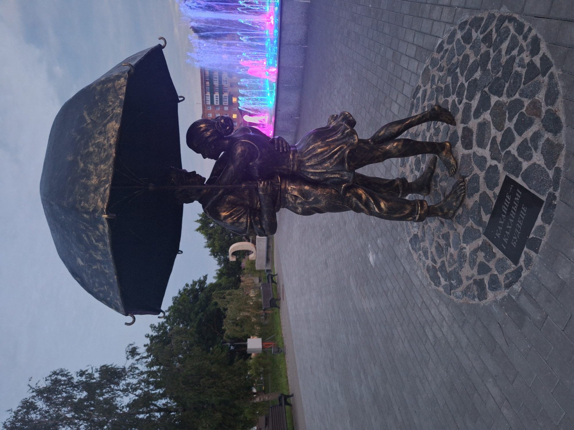 A bronze statue of a couple dancing under an umbrella stands on a paved plaza in Kapyĺ, Belarus. The statue depicts a man and a woman in a close embrace, with the man holding an umbrella over them. The woman wears a flowing dress, and both figures are bare-footed. The statue is positioned on a circular stone base.
In the background, a fountain sprays water illuminated with pink and blue lights. A building with multiple stories and windows is visible behind the fountain. The plaza is paved with gray interlocking bricks, and there are benches and trees on the left side of the image. The sky is overcast, suggesting it might be dusk or a cloudy day. A plaque with text in an unfamiliar alphabet is visible at the base of the statue.