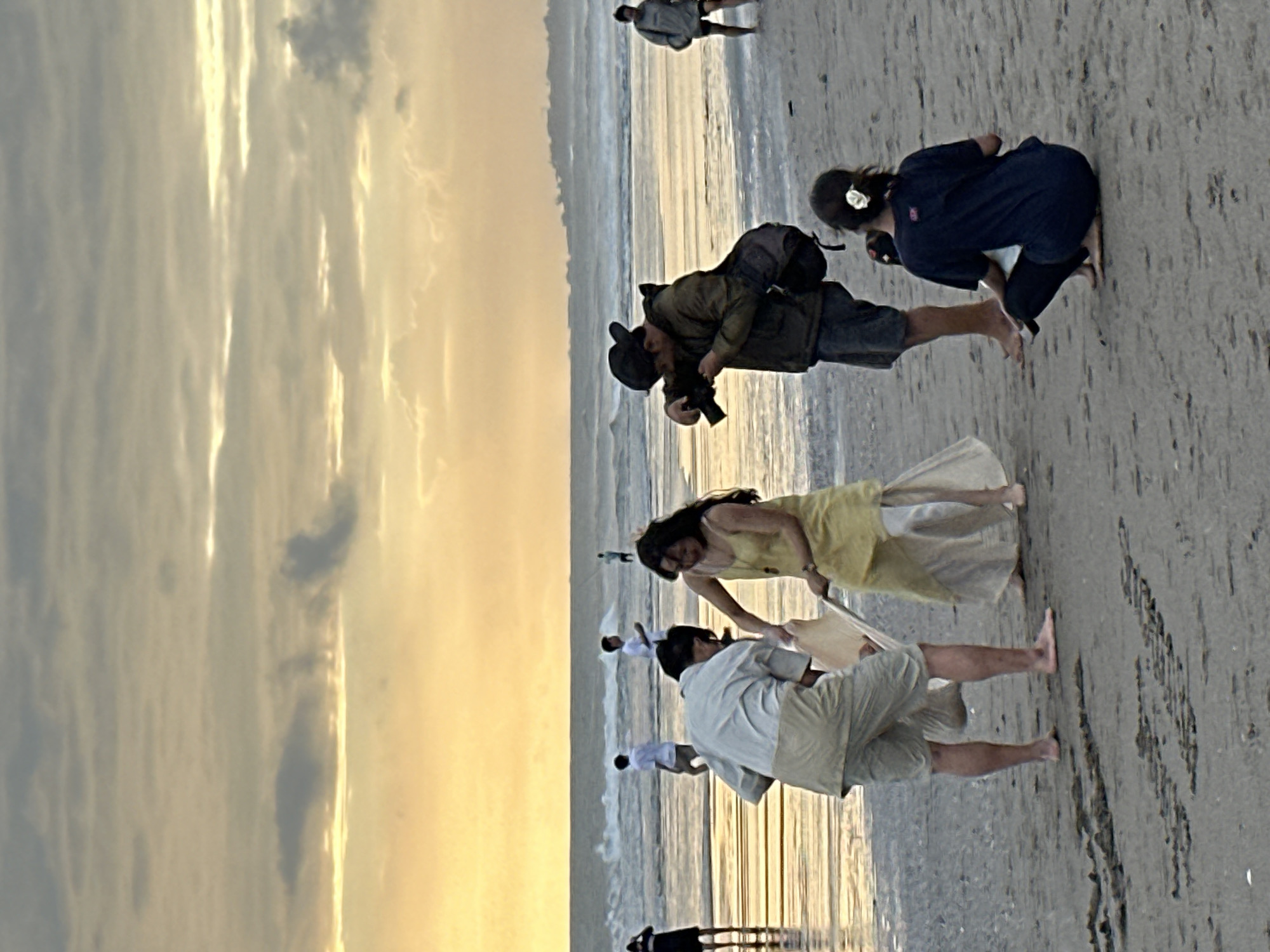 The image depicts a beach scene in North Kuta, Indonesia, likely during sunset. In the foreground, two individuals are interacting, with one appearing to be a photographer with a camera and a bag slung over his shoulder, focused on the other individual who is dressed in a yellow garment. Another person in a light-colored shirt and shorts is partially visible, facing away from the camera and interacting with the person in yellow. Further back, several other individuals are present on the beach and in the shallow water, some walking and others standing. The ocean is visible with waves breaking near the shore, and the sky is filled with dramatic, clouded formations illuminated by a warm, golden light, indicative of a sunset or sunrise. The sand on the beach shows some texture and footprints.