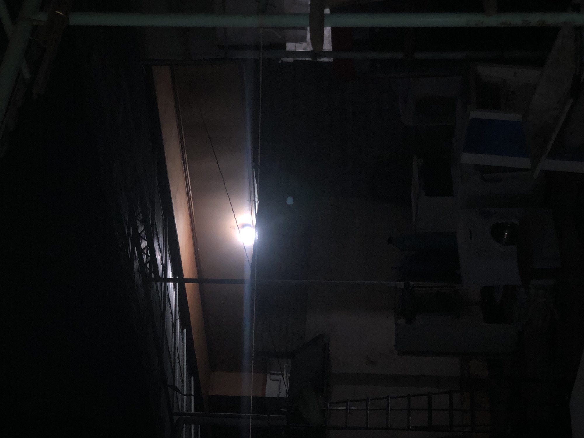 The image depicts an interior space, likely an industrial or storage area, at night. A single, bright light bulb hangs from the ceiling, casting a strong glow and creating a significant lens flare. This light illuminates a portion of the scene, highlighting a vertical beam and some metallic structures. The rest of the space is shrouded in darkness.
The setting appears somewhat cluttered, with various objects and structures visible in the dim light. To the left, a dark, shadowy area suggests a large, open space or a wall covered in some material. A metal grate or fence is visible in the foreground. Towards the right, hints of what could be machinery or large appliances are discernible, some appearing white and blue, possibly representing storage units or industrial equipment. Overhead, pipes and conduits run across the ceiling.
The overall impression is one of an abandoned or disused space, with the stark lighting emphasizing its emptiness and perhaps a sense of neglect. The time of day is clearly night due to the artificial light source and the darkness. There are no people visible in the frame, and no specific events are taking place beyond the ambient lighting. No text is visible. The scene provides a stark and somewhat unsettling atmosphere due to the strong contrast between light and shadow.