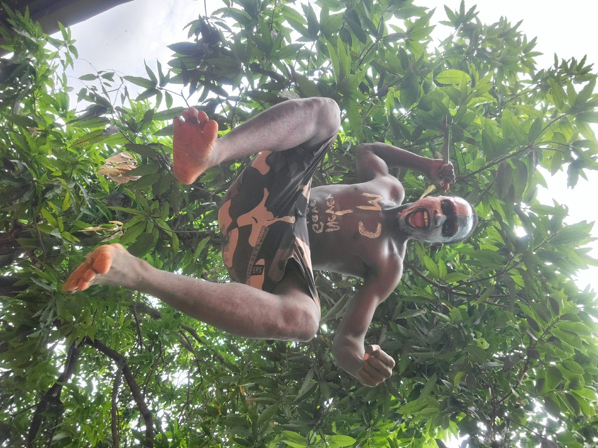 A low-angle outdoor shot depicts a man hanging upside down from a tree. The man is shirtless and wearing camouflage shorts. He has paint on his face, with white around his eyes and red around his mouth. There appears to be writing in white paint on his torso. He is grasping onto the tree branches with his hands. The leaves of the tree create a dense canopy around him. The location is Onitsha, Nigeria, indicated by the presence of Ijele masquerade paintings on the man’s face and body.
