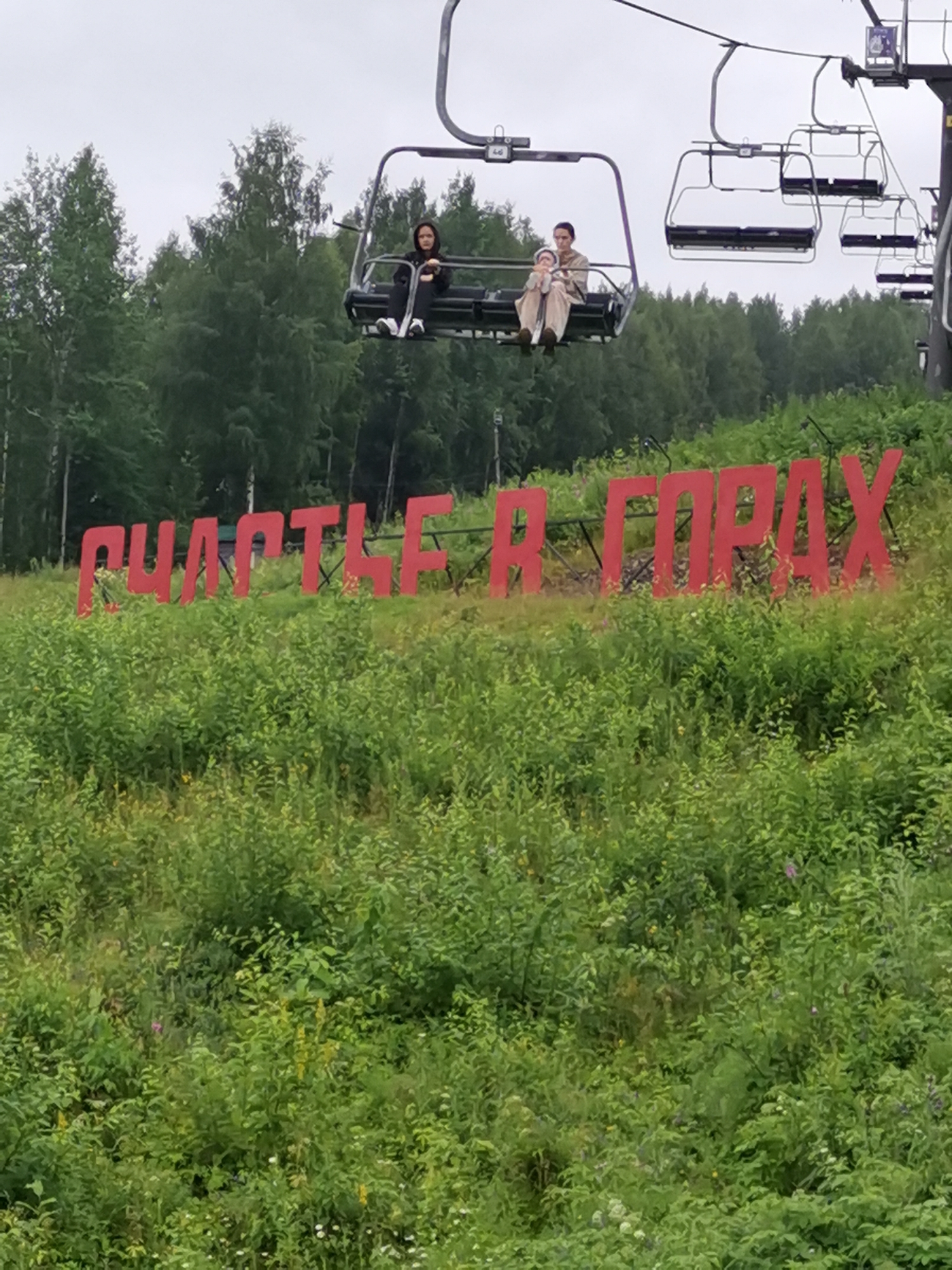 The image shows a ski lift with several empty chairs and one chair occupied by two people and a baby. A person in a black hooded sweatshirt is in one chair, and another person in a light-colored outfit is holding a baby in a subsequent chair. In the background, there is a sign with large red letters that spell "СЧАСТЬЕ В ГОРАХ" which translates to "HAPPINESS IN THE MOUNTAINS." The sign is positioned on a grassy slope with trees in the background, suggesting a mountainous or hilly landscape. The overall environment appears to be a recreational area, likely a ski resort, in Gubakha, Russia, given the textual content of the sign. The sky is overcast, indicating a cloudy day.