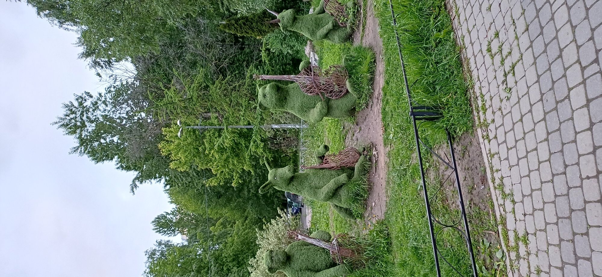 A line of topiary animals, likely rabbits, with instruments are placed along a path in an urban park in Unknown, Unknown. Each animal has a wire-framed instrument; the animals and instruments are made of green foliage. The animals are positioned on a dirt path with green grass on either side. A decorative black metal fence runs alongside the path. To the right is a paved walkway with grey cobblestones. Lush green trees and vegetation fill the background, and a street light is visible among the trees. The sky is overcast.