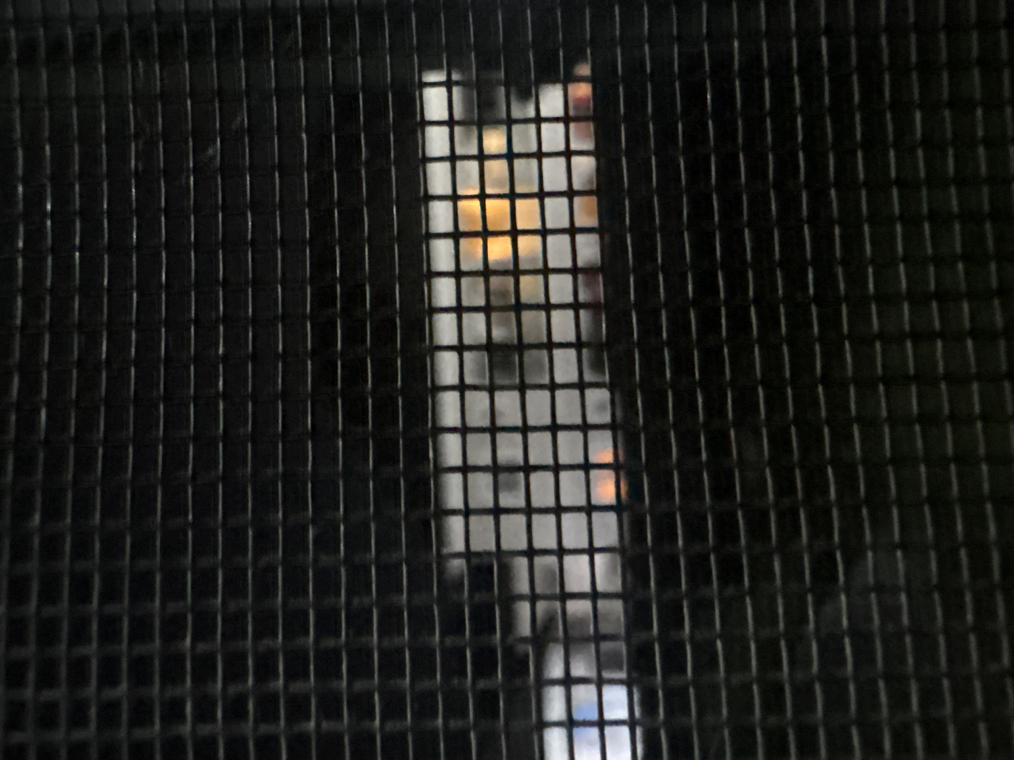 The image displays a close-up view of a black metal grill, obscuring a scene behind it. Through the mesh, a dimly lit interior or outdoor area can be partially discerned. There are faint hints of white structures and a bright orange-yellow light source, suggesting artificial lighting or a flame. The lighting is inconsistent, with some areas appearing brighter than others. The overall impression is that of looking through a barrier at a distance. The location in Angwan Sayawa, Nigeria, is not visually discernible from the provided image.