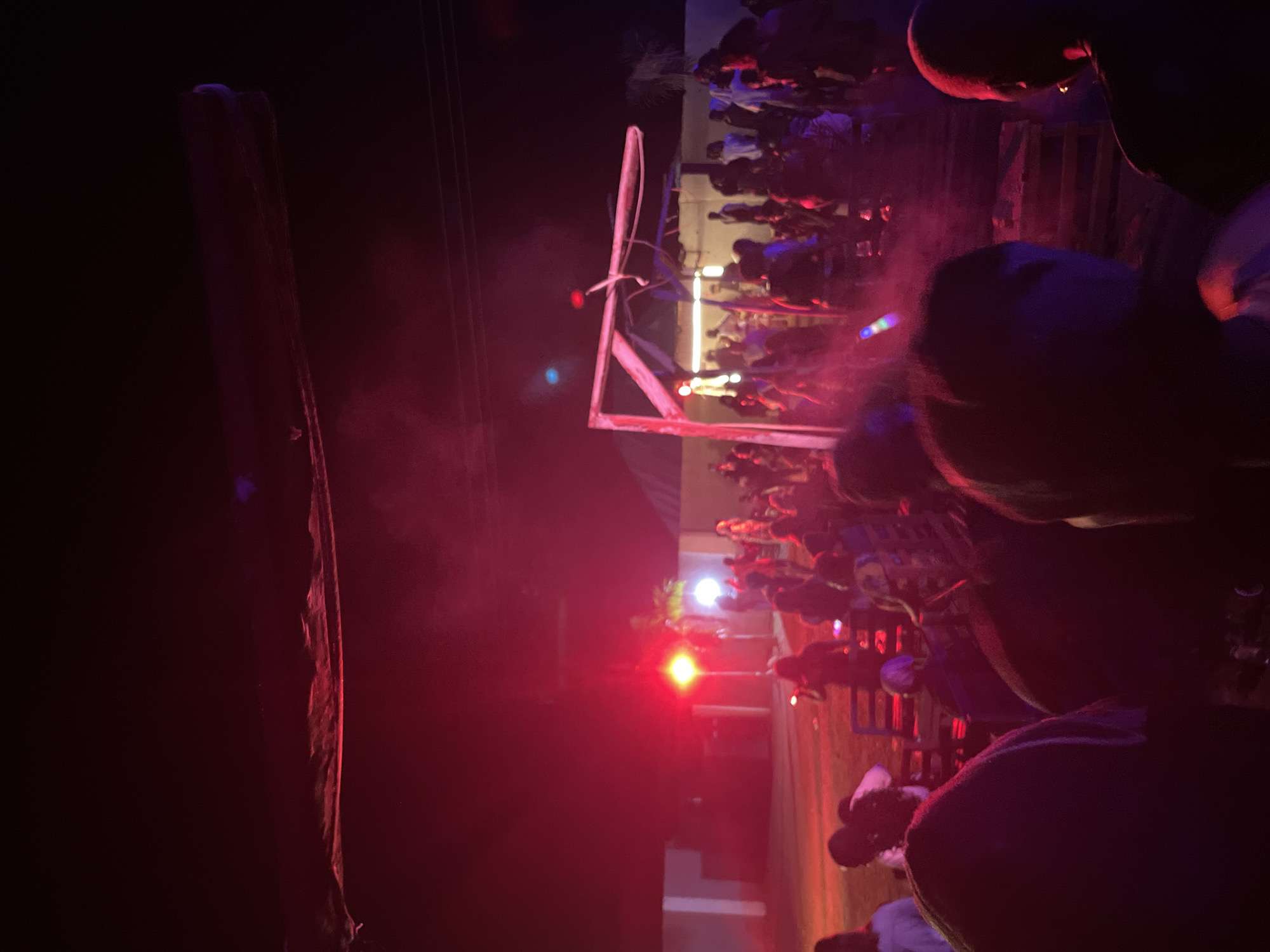 The image depicts an outdoor gathering at night, illuminated by red and white lights, in Durumi, Nigeria. A crowd of people, appearing to be mostly young adults, are present, some standing and others seated on makeshift benches or pallets. In the background, there is a structure resembling a small stage or platform with a wooden frame. Red lights are prominently cast on the scene, creating a hazy atmosphere. The overall impression is of a lively social event or celebration.