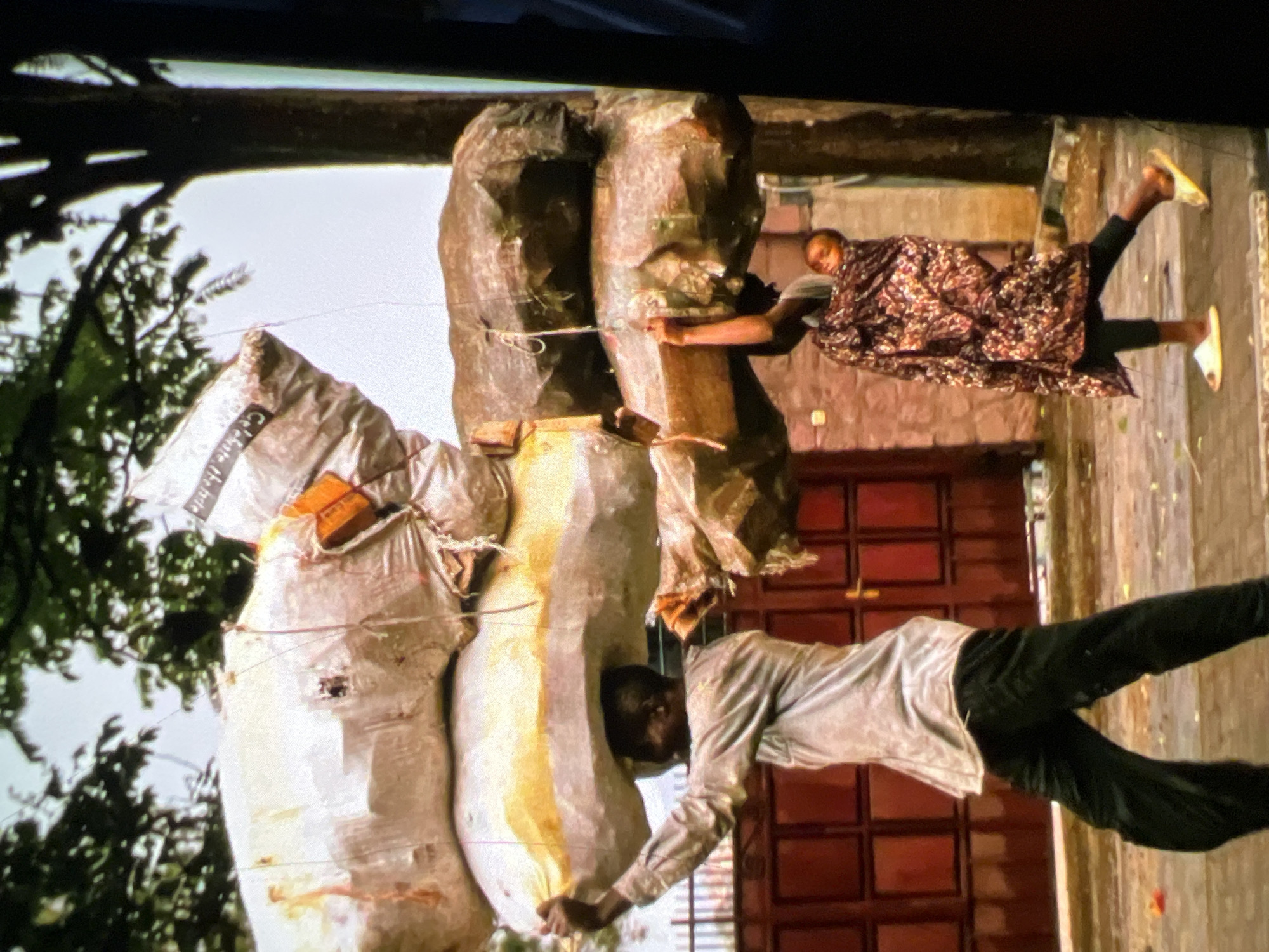 In Lagos, Nigeria, two individuals carry heavy loads balanced on their heads. A man in a light-colored shirt and dark pants balances a stack of at least three large, irregularly shaped sacks, one white and two darker, on his head. To his right, a smaller individual, possibly a child, wrapped in a patterned cloth and wearing slippers, supports a portion of the load with their hands. They are walking on a paved surface, with a red double gate and a tree in the background. The scene appears to be outdoors, with overcast skies.