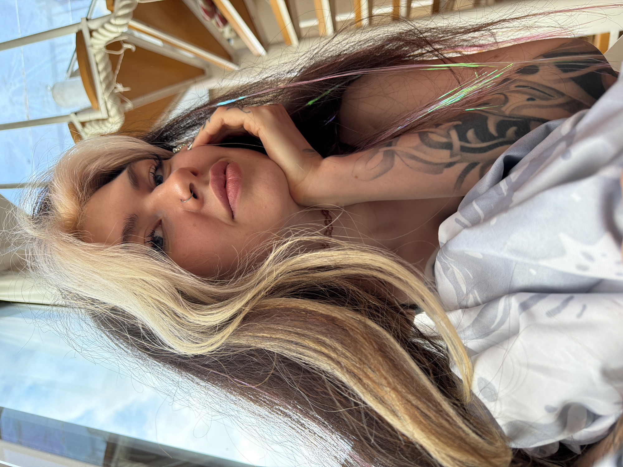 A young woman with blonde and brown hair poses for a photograph. She is wearing a nose ring, and her right arm is tattooed. She is lying on a surface covered with a grey and white patterned fabric. In the background, a blue sky is visible through a window. A white spiral staircase is also visible. Based on the architecture and the potential proximity to the sea, the location may be Budva, Montenegro.