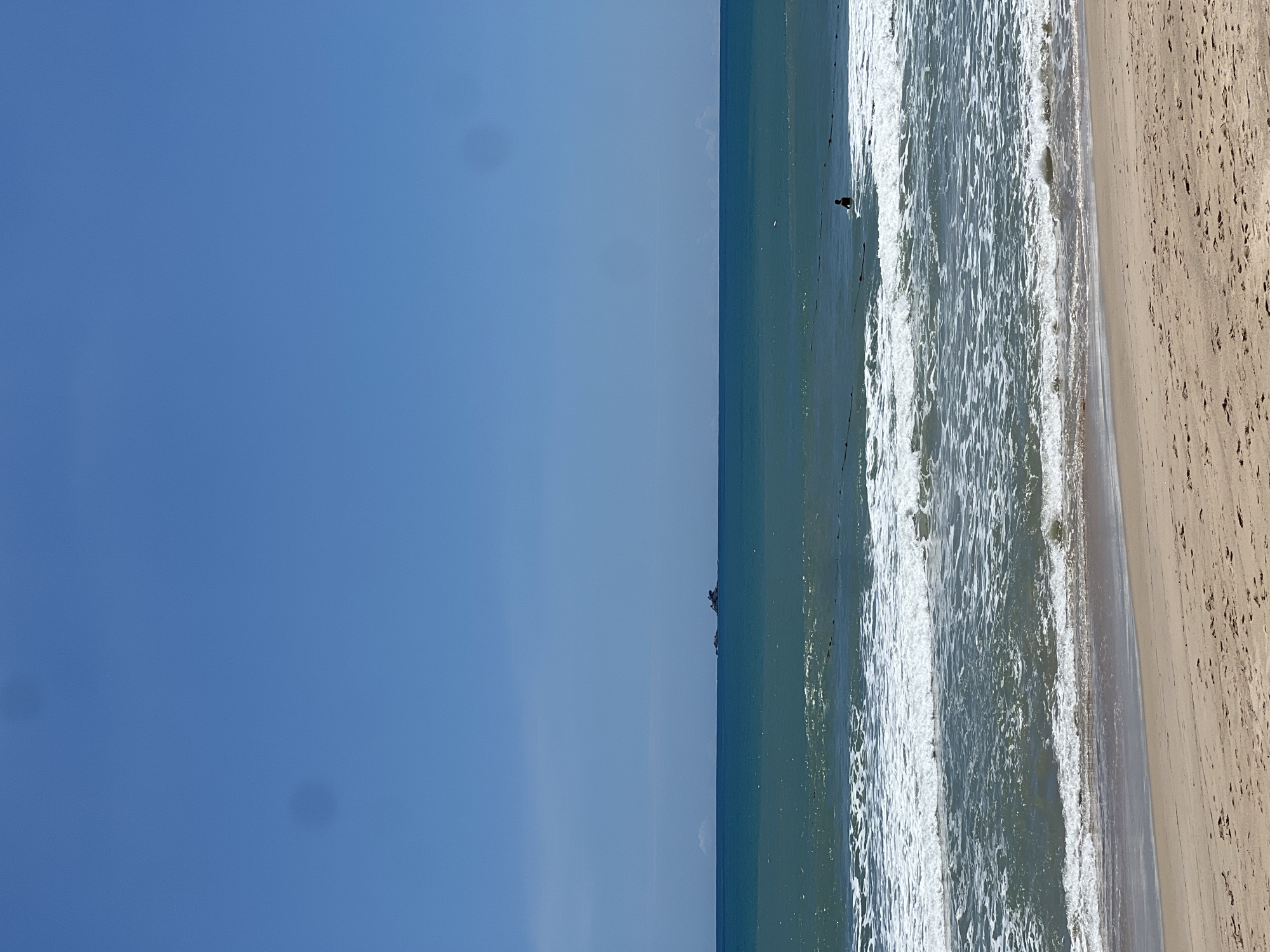 A wide shot captures a serene beach scene under a clear blue sky. The sandy shore occupies the bottom portion of the frame, showing subtle footprints. The ocean stretches across the middle ground, with gentle waves creating white foam as they approach the shore. In the distance, a lone figure is visible in the water, appearing to be swimming or wading. Further out at sea, a small island with rocks and sparse vegetation is discernible on the horizon. The overall impression is one of tranquility and natural beauty.