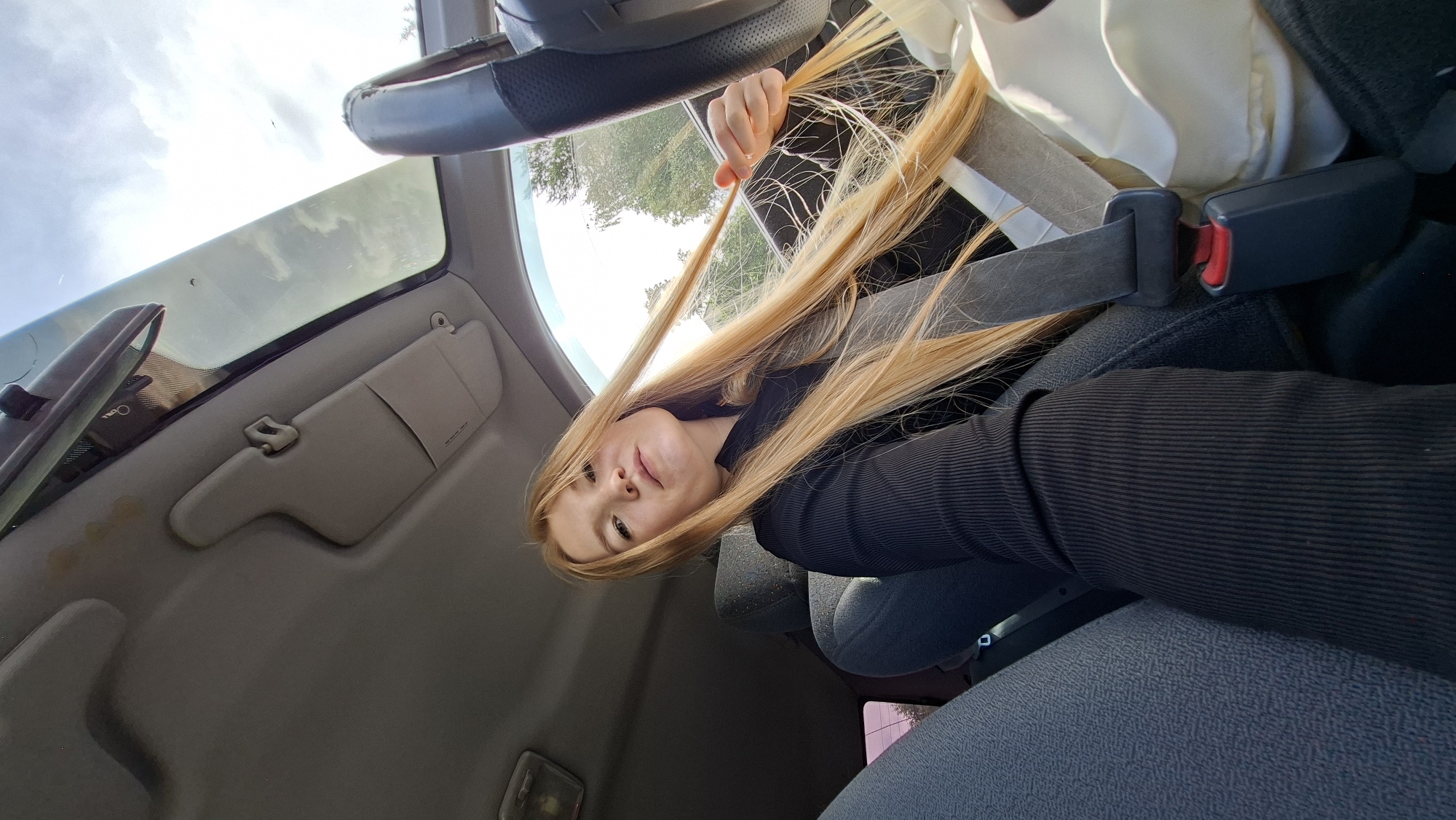A young woman with long, blonde hair is sitting in the driver's seat of a car. She is wearing a black, ribbed long-sleeved shirt. Her right hand is holding a section of her hair. The car has a sunroof that is open, and the sky with clouds is visible through it. The interior of the car, including the dashboard, sun visor, rearview mirror, and car seat, is visible. The woman is looking towards the camera with a neutral expression. The scene appears to be set during the daytime, with natural light illuminating the interior of the car. The woman is likely in the city of Chaikovsky, Russia, given the context provided.