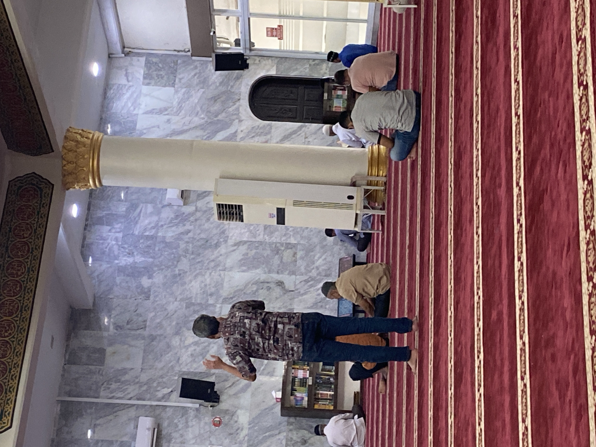 The image depicts the interior of a mosque in Lhokseumawe, Indonesia, likely during prayer. Several men are seated on a red carpet with gold patterned lines. In the foreground, a man stands facing away from the camera, with his hands raised. In the middle ground, other men are seated, some appearing to be in prayer or contemplation. A large white column with a gold capital stands prominently, dividing the space. The walls are marble-patterned, and a bookshelf is visible on the left. An air conditioning unit is mounted on the wall, and speakers are positioned high on the wall. The overall lighting suggests an indoor environment.
