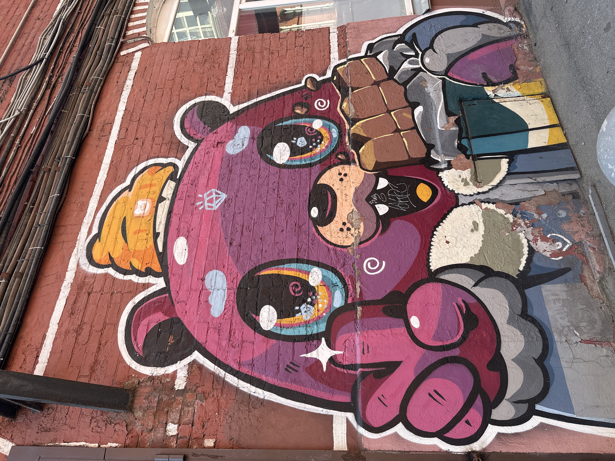 The image displays a vibrant graffiti mural of a cartoon bear on a weathered red brick wall. The bear is predominantly purple and pink, adorned with an orange hat and large, expressive eyes. It is holding a large piece of chocolate and making a peace sign with its left hand. The mural also features smaller details like clouds, a diamond, and swirling patterns. Below the bear, a section of the wall is painted with colorful geometric shapes, partially covering a metal hatch. The environment appears to be an urban setting, possibly an alleyway or side street, with a window visible on the right side of the wall and overhead electrical wires running along the top. The ground in front of the mural is asphalt, with a concrete curb and a large, spherical object partially visible in the foreground. The style of the graffiti is bold and graphic, typical of street art. The location is identified as Moscow, Russia.