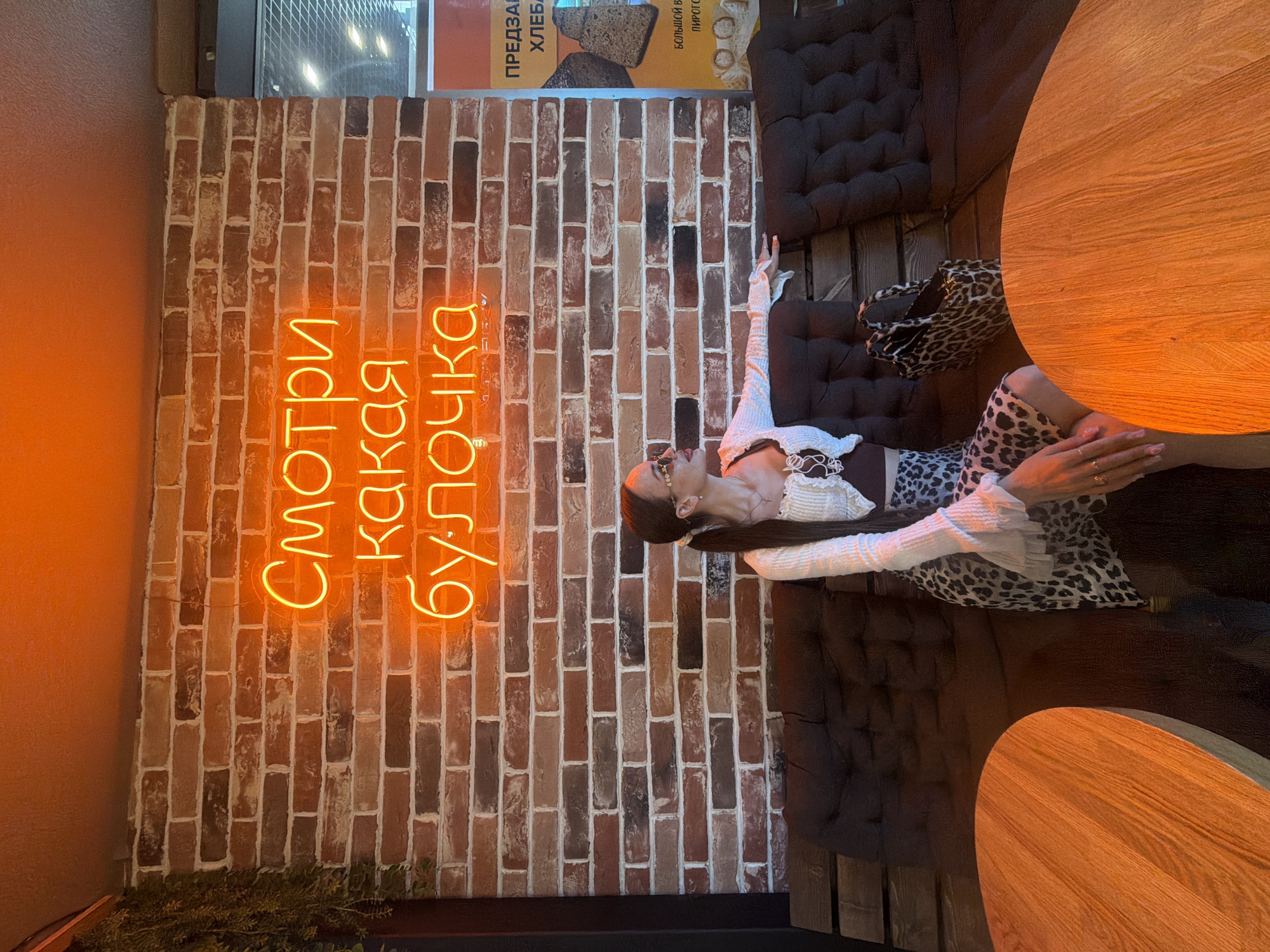 An indoor scene within a cafe or bakery in Saint Petersburg, Russia, features a young woman seated on a dark brown, tufted bench against a rustic brick wall. The woman has long, dark brown hair in a ponytail and wears round sunglasses, a white ribbed cropped cardigan over a dark top, and a leopard-print midi skirt. She has multiple rings and a cross pendant necklace. Her left arm rests on the bench's back as she looks to her right, away from the camera. A leopard-print tote bag is positioned beside her on the bench. Above her, an orange neon sign on the brick wall displays the Russian phrase "Смотри какая булочка" (Look what a bun/cutie) across three lines. To the far left, green foliage hangs vertically. In the foreground, two empty, round wooden tables are partially visible on either side of the woman. On the far right, a yellow and blue poster advertises "ПРЕДЗАКАЗ ХЛЕБА" (Pre-order bread) and "БОЛЬШОЙ ВЫБОР ПИРОГОВ" (Large selection of pies) in Russian, with images of bread. The environment is warmly lit.