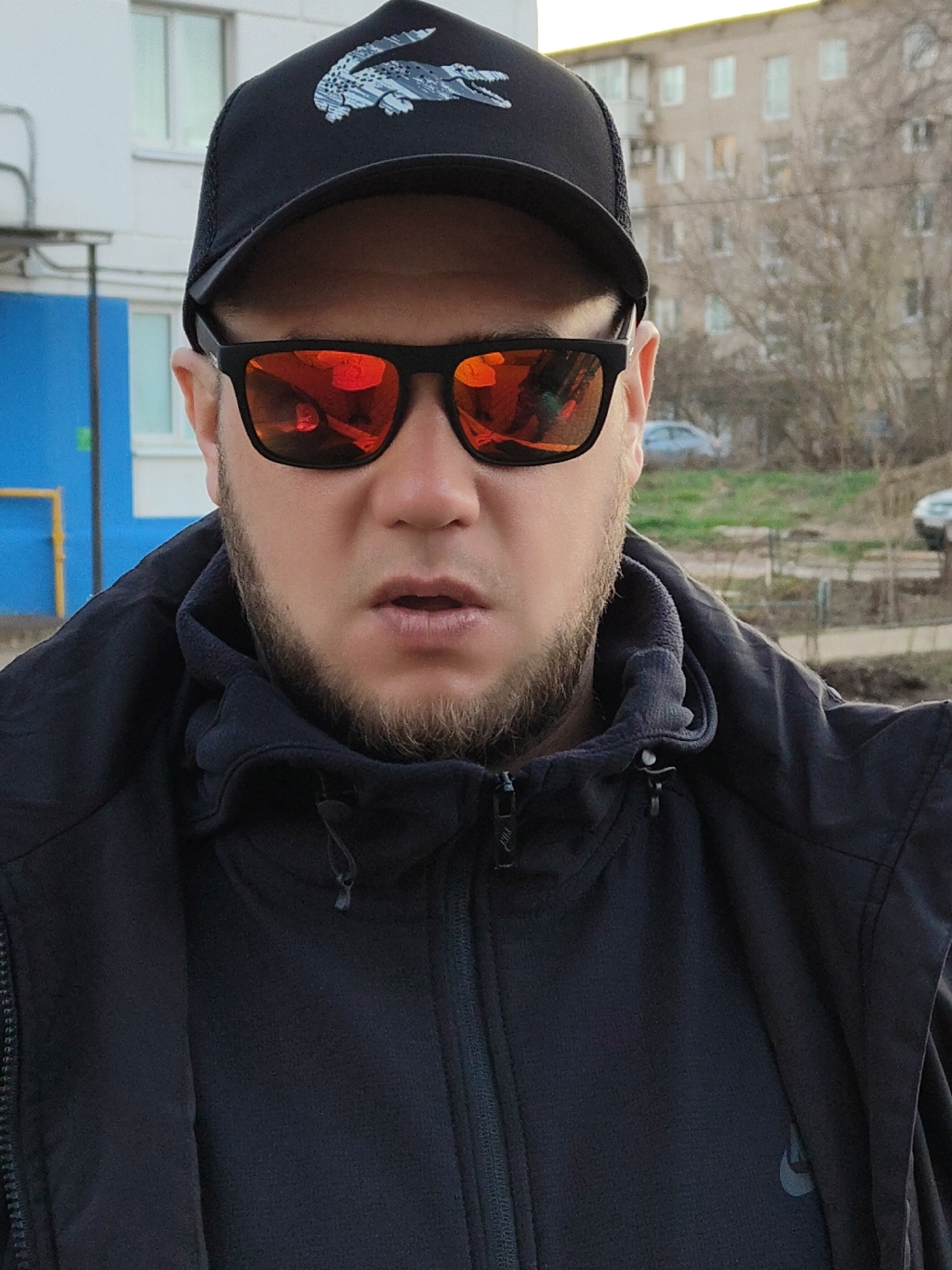 A man is depicted in a mid-shot, facing the camera directly, against an urban or suburban backdrop. He has a short, trimmed beard and mustache. He wears a black baseball cap featuring a distinctive white and gray stylized crocodile logo, identifiable as Lacoste. His eyes are obscured by black-framed sunglasses with highly reflective orange-red lenses, which show reflections of the environment.
He is dressed in a black hooded jacket, worn over a dark-colored inner layer, possibly fleece, visible around his neck. A subtle, dark Nike swoosh logo is present on the left chest area of the inner garment. His mouth is slightly open, conveying a neutral or slightly surprised expression.
The background includes parts of residential buildings. To the left, a light-colored building with a prominent blue section and white windows is visible. To the right, another light-colored multi-story building is situated behind bare trees, suggesting a cooler season like late autumn or winter. A car is vaguely visible in the distant background. The lighting is soft, indicative of an overcast day or late afternoon.