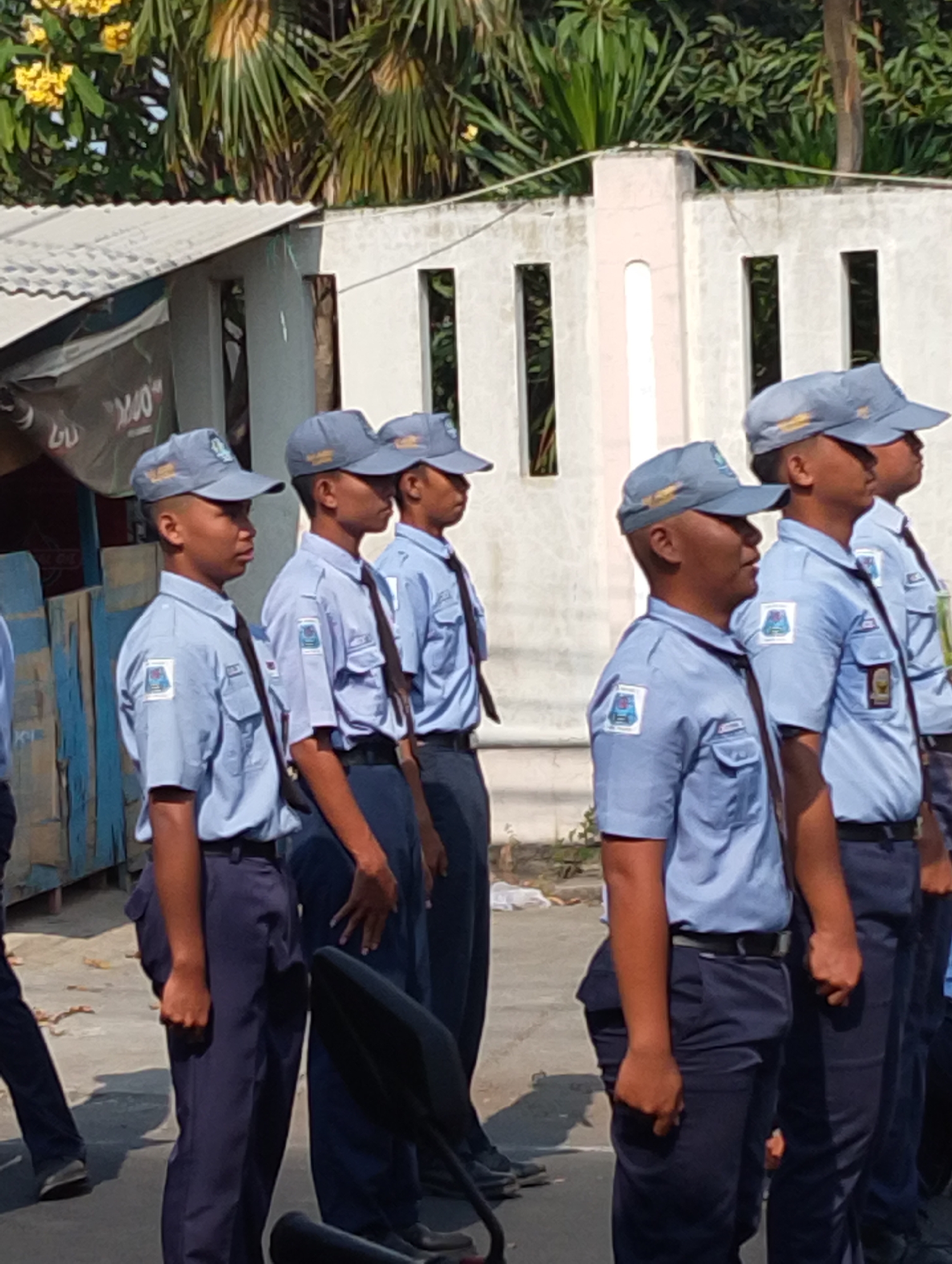 In Manaruwi, Indonesia, a group of five young males, presumably students, stand in formation outdoors. They are dressed in light blue collared shirts with dark ties, dark blue pants, and matching grey caps. Each shirt features a patch on the left breast depicting a blue and white shield with a crest and text. The students appear to be standing at attention, facing to the right of the frame. The background consists of a light-colored wall with rectangular openings, a corrugated roof structure, and lush green foliage with yellow flowers. The ground beneath them is paved.