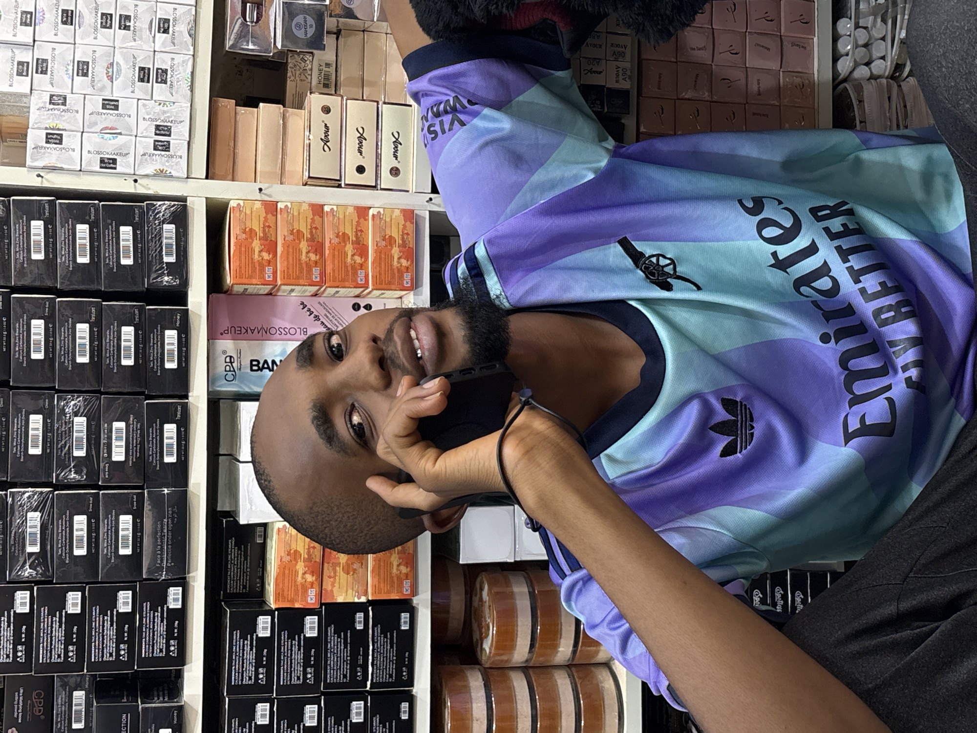 This media file shows a close-up, vertically oriented shot of a young Black man in Jos, Nigeria, holding a smartphone to his ear and looking towards the camera. He is wearing a light blue and purple t-shirt with a camouflage pattern and text that appears to read "Emirates" and "BETTER" in black lettering, along with the Adidas logo. The man has short black hair and a beard.
The background is filled with shelves displaying various cosmetic products, predominantly makeup items in boxes and containers. Many of the boxes have text in English, some of which are identifiable as "BLOSSOMMAKEUP," "Translucent," "Spotless," and "HONEY." Other text includes brand names like "CPD" and "BAN." The abundance of products suggests the setting is a retail store or a market stall specializing in cosmetics. The lighting appears to be artificial, consistent with an indoor setting. The man's expression is neutral, possibly indicating he is engaged in a phone conversation. No specific time of day or weather conditions are discernible.