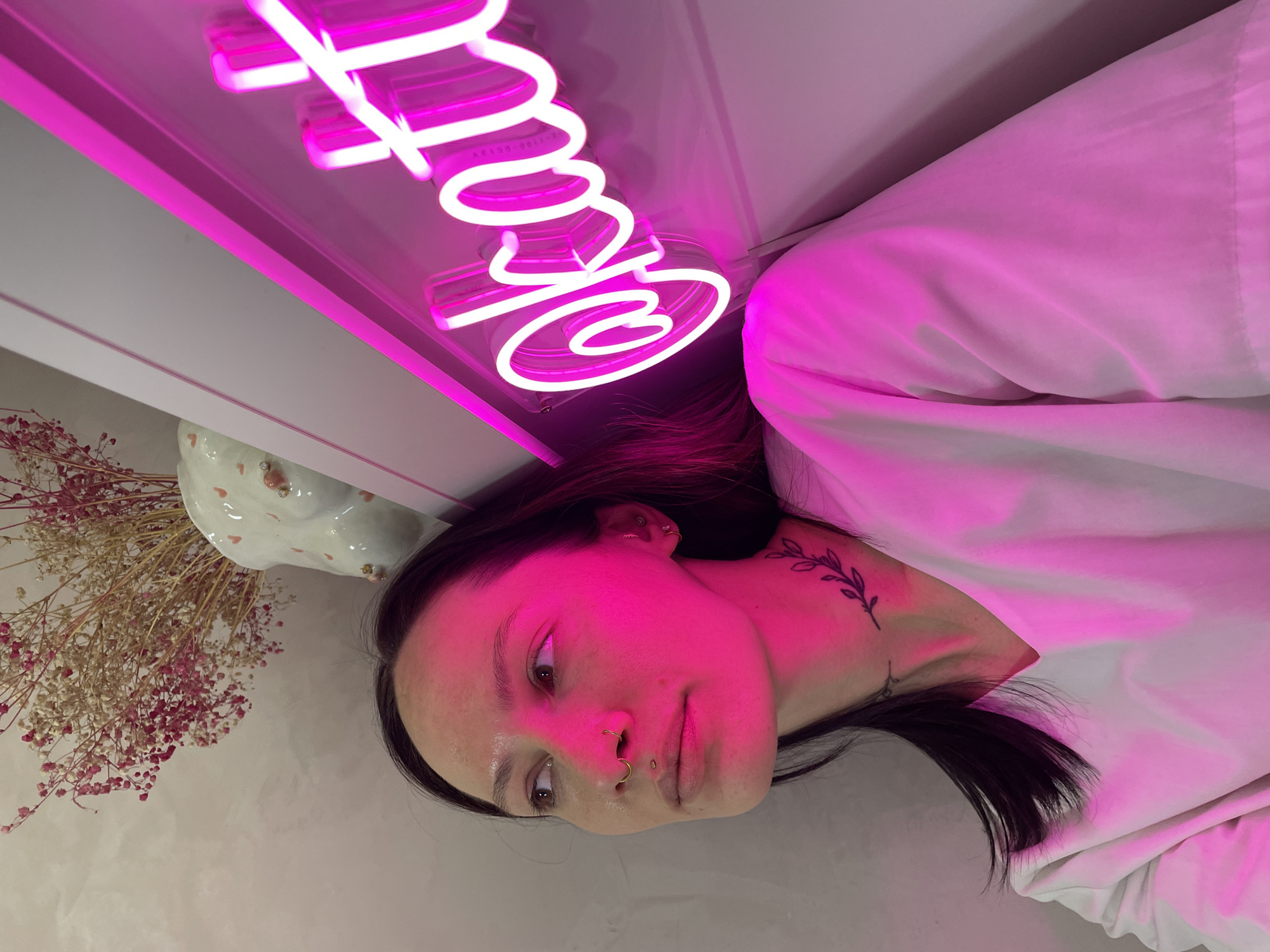 A woman lies on her side with a bright pink neon sign with text above her. She wears a white shirt. She has dark hair and a tattoo on her neck. There is a vase of dried flowers to the left of her head. The location is unknown.