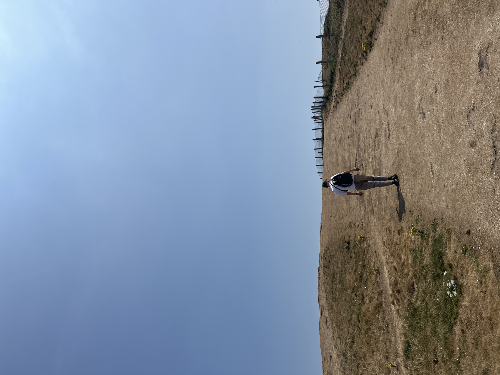 A person walks up a dirt path on a dry, grassy hill under a clear blue sky. The path is wide and leads towards a wooden fence that runs along the top of the hill. The person is wearing a white t-shirt, beige pants, and a black backpack. The grass on the hill is mostly brown and dry, with patches of green and some small yellow and white wildflowers. In the distance, a body of water is visible to the right of the path. The image appears to have been taken on a sunny day. This scene is consistent with the landscape found at Seaford Head, Seaford, United Kingdom, known for its chalk cliffs and coastal paths.
