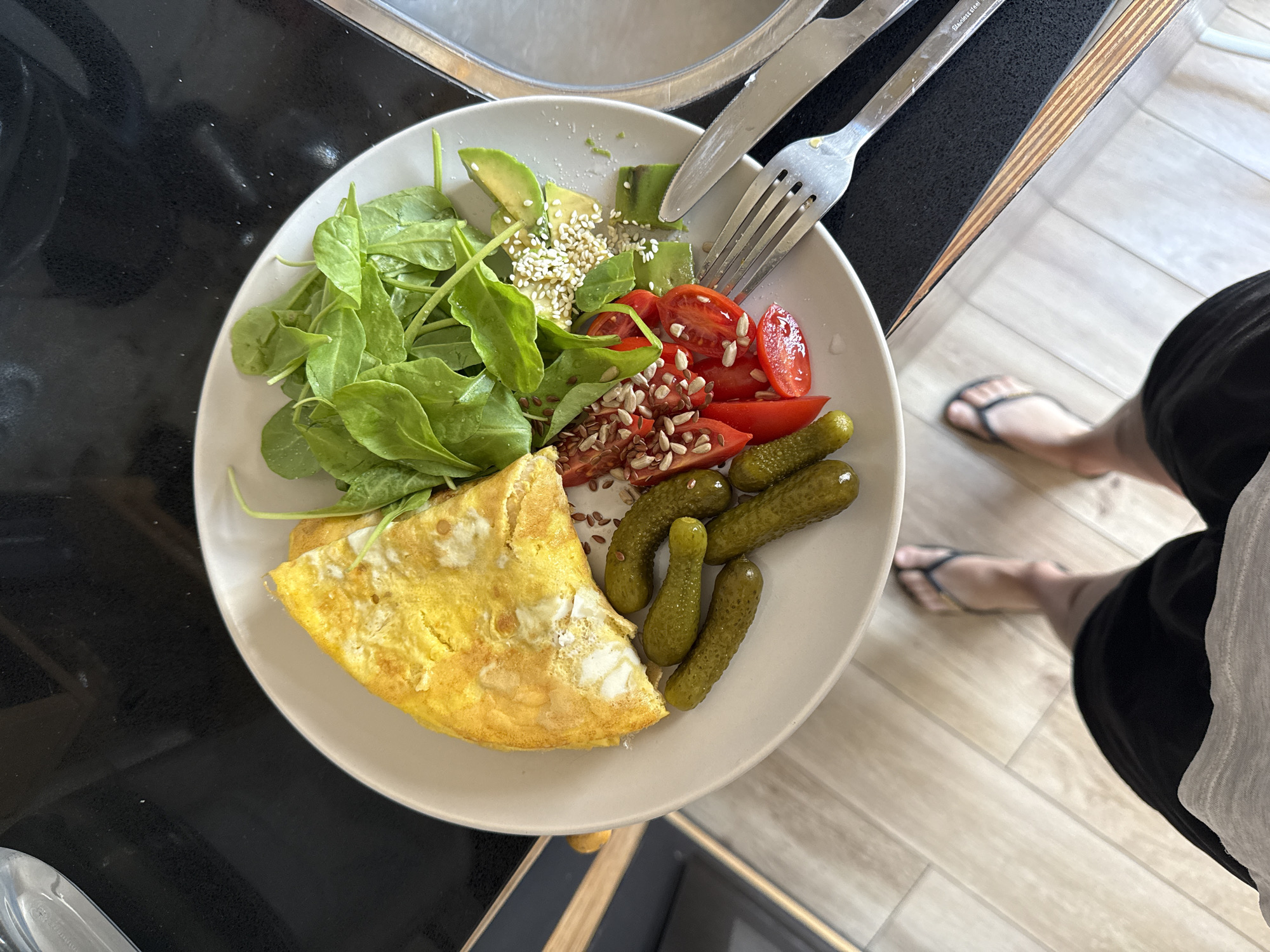 A balanced meal is prominently featured on a light-colored, round plate, positioned on a dark, reflective kitchen counter. The plate holds a folded yellow omelette, a portion of fresh green spinach, sliced avocado adorned with white sesame seeds, and halved red cherry tomatoes sprinkled with sunflower and flax seeds. Several small green gherkin pickles accompany the main components. A silver fork and knife are placed on the plate, with the knife bearing the text "Stainless steel".
In the upper left background, a portion of a stainless steel kitchen sink is visible. To the right of the plate, a person is seen from the waist down, wearing black shorts and black flip-flops, standing on a light-toned wooden or tiled floor. The scene suggests a domestic kitchen setting, possibly during breakfast or brunch due to the meal's composition. The overall impression is one of a freshly prepared, healthy meal ready to be enjoyed. The location context provided is Tel-Aviv, Israel.
