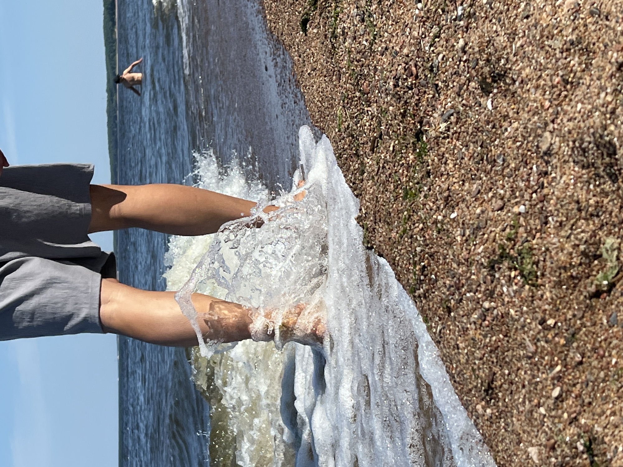 A low-angle, full-shot perspective captures a person's legs and feet submerged in ocean water, with a wave crashing around their ankles. The individual wears gray shorts. In the distance, another person is visible swimming in the ocean. The foreground is dominated by a pebble beach, with the ocean stretching out to the horizon under a clear blue sky.