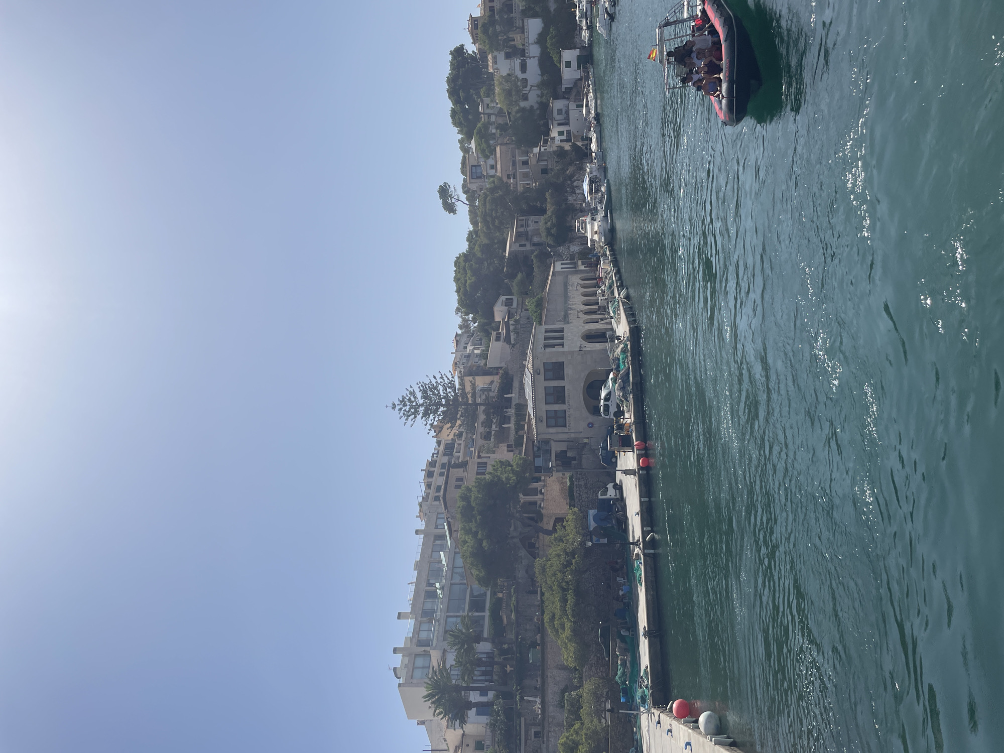 A bright, sunny day illuminates a harbor in Santanyí, Spain, with buildings lining a hillside. In the foreground, a dark, choppy body of water fills the lower portion of the frame. To the right, a rigid inflatable boat with an awning, carrying at least eight people, motors across the water. The boat displays a Spanish flag. Along the left edge, a pier juts out, with red and white buoys. Beyond the pier, a paved walkway borders a line of parked vehicles, including a white van and two SUVs. Fishing nets are draped over the walkway and an adjacent wall. Buildings of varying architectural styles, from modern multi-story structures with balconies to older stone buildings, ascend the hill behind the harbor. Lush green trees and vegetation dot the landscape. The sky is a clear, pale blue.