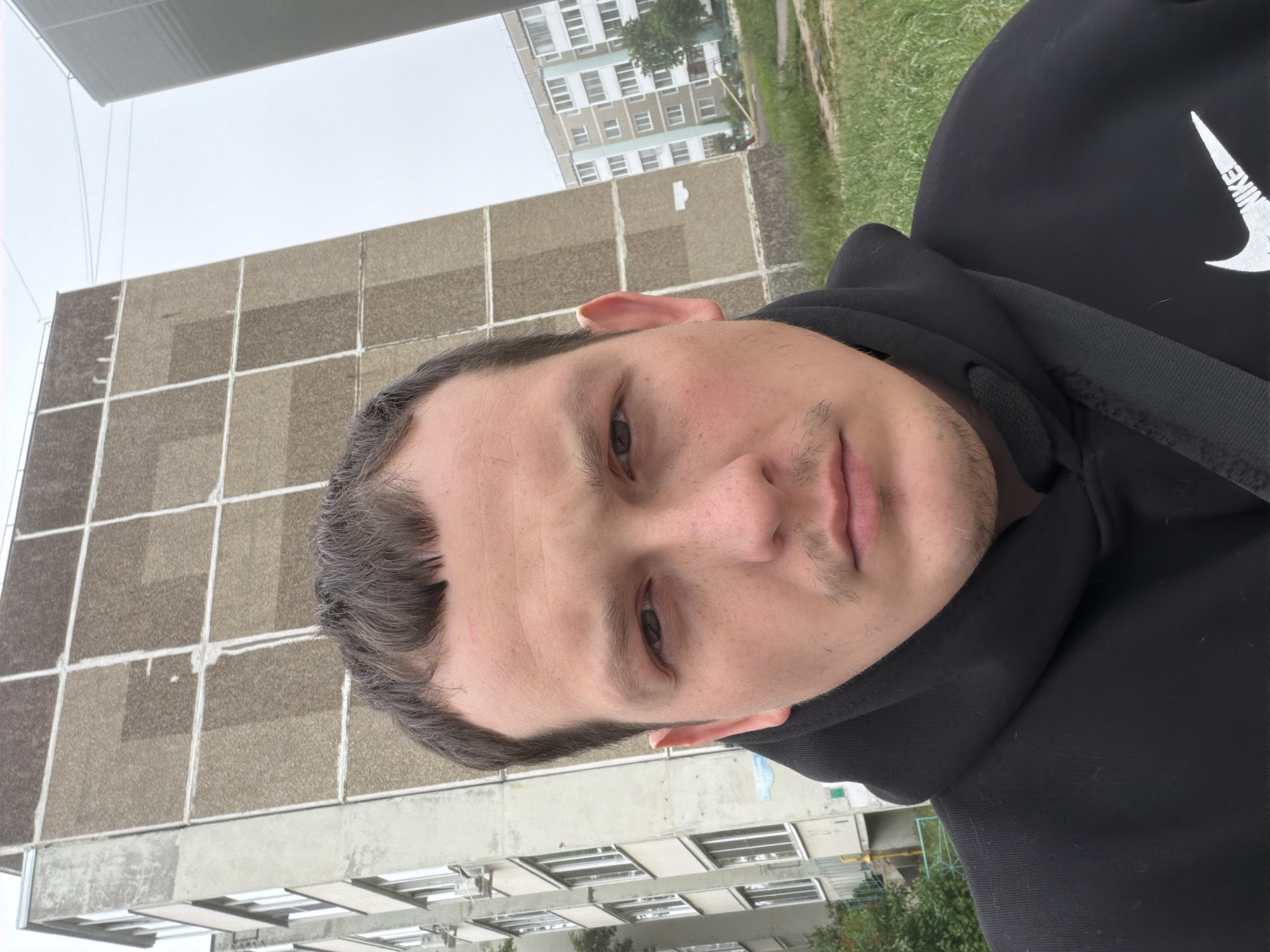 The original image is rotated 90 degrees counter-clockwise, showing a young man in a selfie taken outdoors. The man, positioned in the foreground, has dark hair, dark eyes, and light stubble/mustache, looking directly at the camera with a neutral to slightly pensive expression. He is wearing a black hooded sweatshirt with a prominent white "NIKE" logo visible on his right chest/shoulder. A black strap, likely from a bag, is visible over his right shoulder.
The background reveals an urban or suburban setting, consistent with Chaikovsky, Russia. Two multi-story residential buildings are visible under an overcast sky. The closer building, to the man's left (upper right in the original orientation), is a light brown or grey structure with a grid-like pattern on its facade, suggesting panel construction common in Soviet-era architecture. Further in the background, another taller, lighter-colored apartment building with multiple windows can be seen. A patch of green grass separates the buildings. The sky is uniformly grey and cloudy, indicating an overcast day.