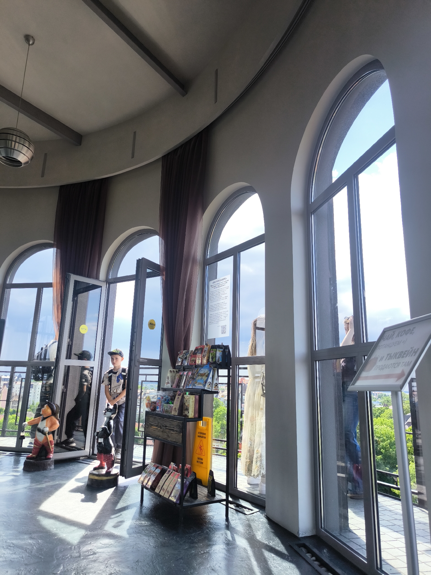 The image depicts the interior of a circular or semi-circular observation area, likely within the Water Tower in Zelenogradsk, Russia, characterized by its curved walls and numerous large arched windows. The flooring is a dark, textured material, and the ceiling is light-colored with dark linear architectural details and a single metallic spherical pendant light. Dark brown curtains are drawn back at several window sections.
Through the clear glass windows, a bright daytime view of a cityscape with extensive green foliage is visible under a blue sky with scattered white clouds. A balcony or terrace with railings runs along the exterior of the building.
Several people are present:
* In the center-left, a young boy wearing a patterned t-shirt, dark pants, and a baseball cap stands near an open glass door, looking towards the viewer. His reflection is visible in the glass.
* To his left, a man in dark attire and a baseball cap is partially visible, positioned near another glass door and looking outwards.
* On the far right, the reflection of a woman with long light-colored hair and a dark top is seen in the window.
* Partially visible on the center-right, a woman's lower body in a long, light-colored patterned dress stands behind a glass pane.
In the foreground, on the dark floor, are two colorful, stylized figurines:
* On the left, a stout figure in an orange jacket, white shirt, and red boots holds a stick.
* To its right, a dark animal-like figure in a red vest and black hat stands upright, holding an object.
Further into the room, on the center-right, a dark wooden display stand with multiple shelves holds various merchandise, likely souvenirs. Adjacent to this stand is a yellow A-frame "CAUTION WET FLOOR" sign, displaying text in both Russian ("ОСТОРОЖНО МОКРЫЙ ПОЛ") and English. On the far right, a silver-framed sign on a stand indicates in Russian, "ЧАЙ, КОФЕ И ТЫКВЕЙН ПРОДАЮТСЯ ТАМ" (Tea, Coffee, and Pumpkin Wine sold there), with a red arrow pointing right. Various informational posters or notices, some with QR codes, are affixed to the glass surfaces of the windows and doors. Sunlight streams through the windows, casting distinct shadows across the floor.