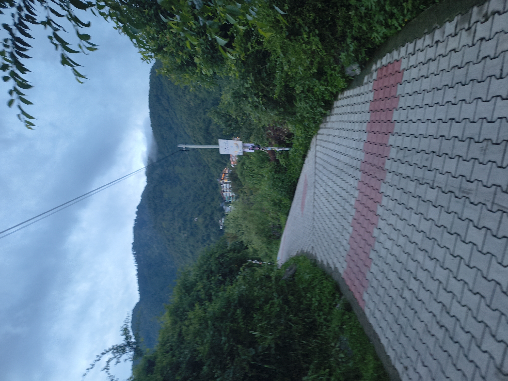 An upward-angled, medium shot captures a paved pathway winding through lush green foliage towards a tree-covered mountainside under a cloudy, overcast sky. The pathway is constructed from interlocking grey pavers with a central strip of red pavers. A tall white utility pole with some attached signage stands to the right of the pathway. Buildings are visible nestled on the mountainside in the distance. The scene conveys a sense of natural beauty and a tranquil, perhaps secluded, location.