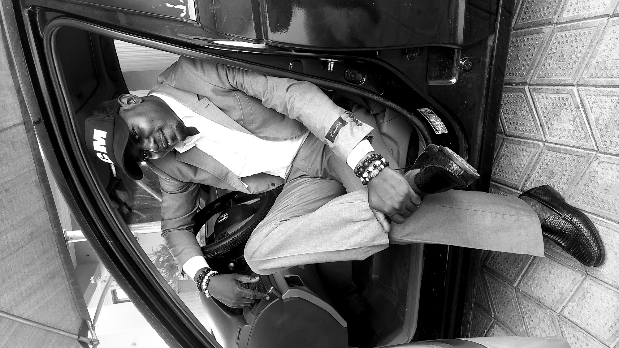 A black and white image captures a stylish man posing horizontally inside a car, looking directly at the camera with a confident, slight smile. He is dressed in a light-colored suit, comprising a jacket with a "Z.ZARA" label visible on the sleeve and matching pants, paired with a white collared shirt. He wears a black baseball cap with "GM" prominently displayed on the front. Both wrists are adorned with multiple beaded bracelets in dark and light tones.
The man's left hand rests on the steering wheel, which features an "H" logo, suggesting a Honda vehicle. His right hand holds his left pant leg. He is wearing black, textured loafers. The car's interior is visible, including the dark dashboard and door frame. Outside the car, on what appears to be the ground or pavement, there are distinctive patterned hexagonal tiles. The scene is well-lit, suggesting it is daytime. The man appears relaxed and fashion-conscious in this setting, located in Sabon Tasha, Nigeria.