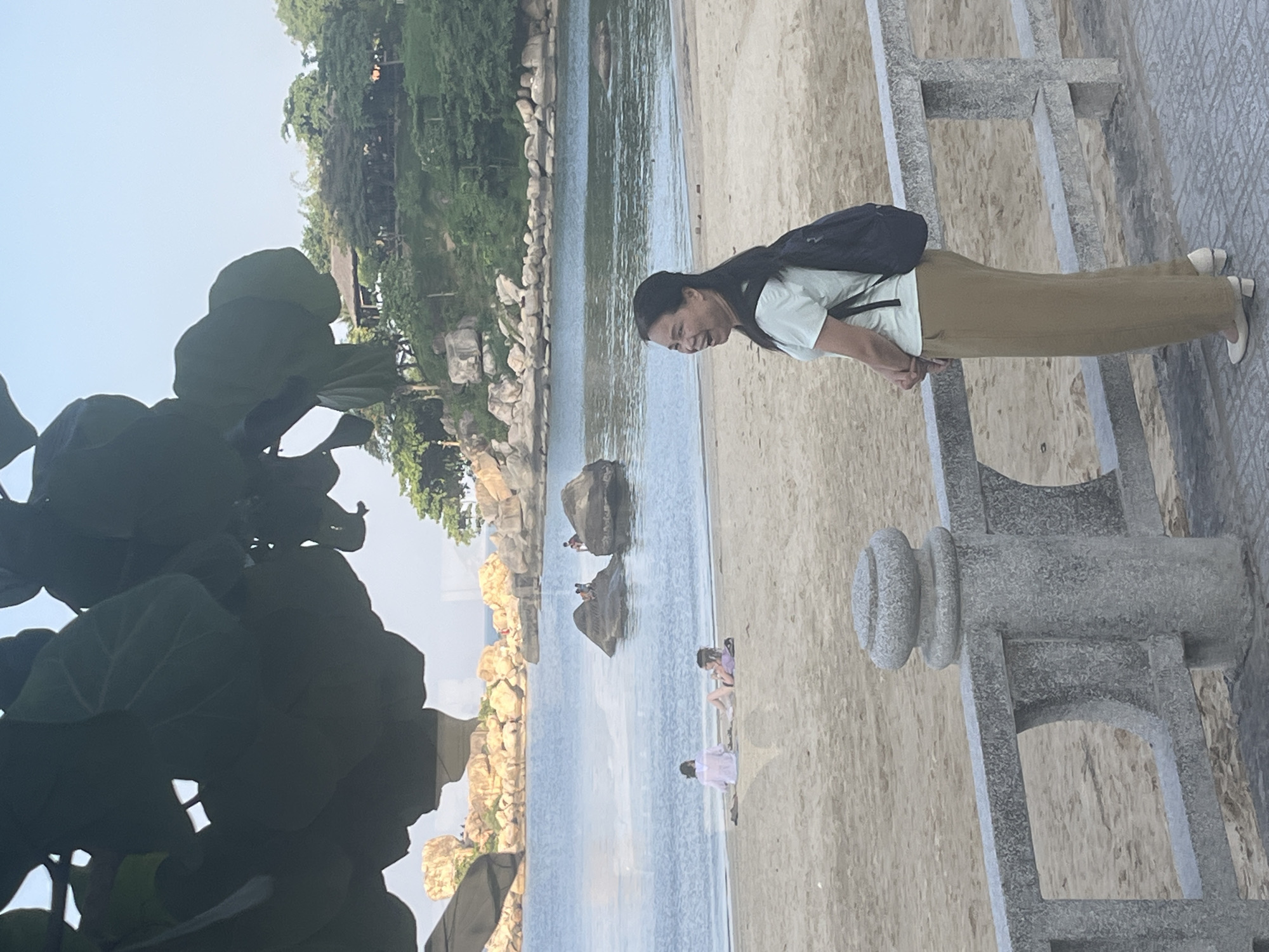 In Nha Trang, Vietnam, a woman with dark hair stands on a paved walkway overlooking a sandy beach and the sea. She is wearing a light green t-shirt, tan pants, and cream-colored heels, and has a dark backpack on. She is smiling and looking to her left. Behind her, the beach is populated by a few individuals. Two people are sitting on the sand near the water's edge, one in a white shirt and the other in a purple top. Further out in the water, several people are gathered on and around large rock formations. The background features a lush, tree-covered hillside with a building structure visible on top. The image is framed by large, dark green leaves from a tree in the foreground.