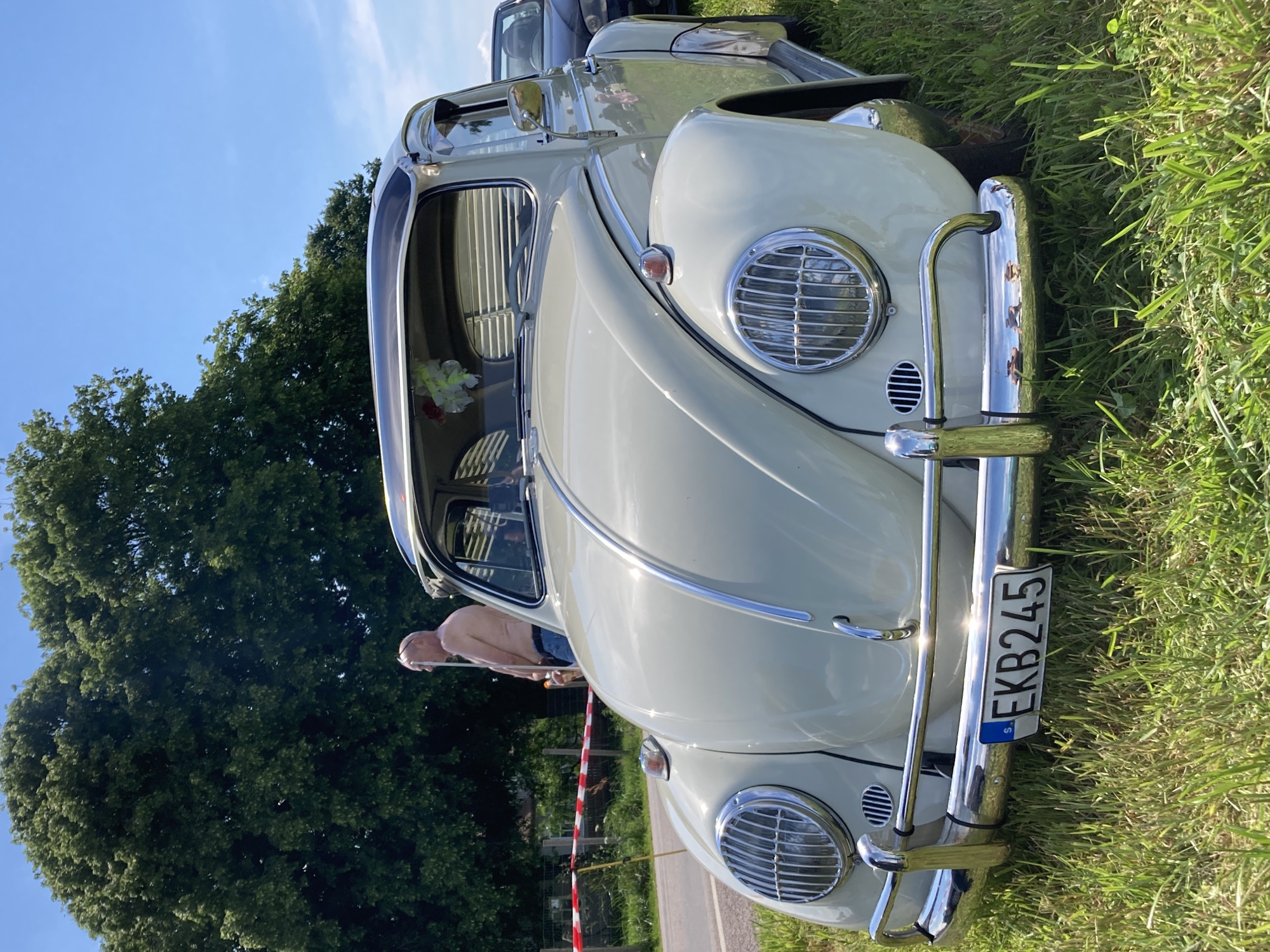 A vintage light cream-colored Volkswagen Beetle is parked on a grassy area next to a road. The car faces the camera, with its chrome bumper and headlights prominently displayed. A license plate with the letters "EKB245" is affixed to the front bumper. In the background, a shirtless man with sunglasses stands near a red and white striped tape barrier. Tall, green trees form a backdrop against a clear blue sky. The image appears to have been taken in Edenberga, Sweden, as indicated by the license plate.