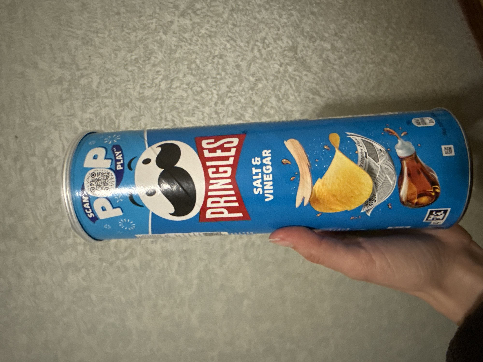 A hand holds a blue cylindrical container of Pringles Salt & Vinegar crisps. The backdrop is a textured, light gray wall. No indication of Saint Petersburg, Russia.