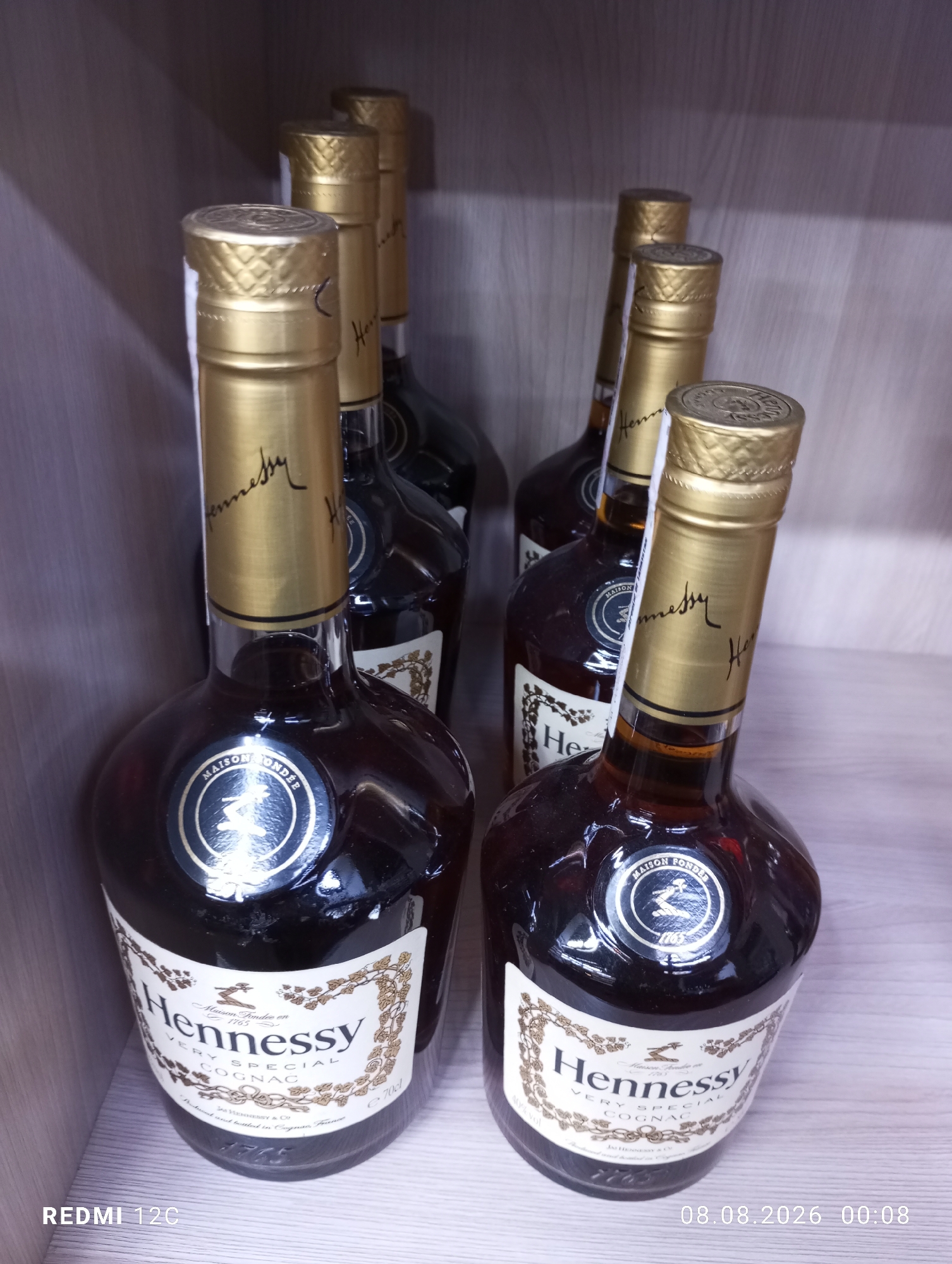 The image shows a collection of Hennessy Very Special Cognac bottles. There are at least six bottles visible, arranged on a shelf. The bottles are dark glass with gold foil caps and labels. The labels are white with gold lettering and intricate designs, featuring the Hennessy logo and text. The bottles are positioned in a way that some are partially obscured by others. The shelf appears to be made of light-colored wood. The background is a dark, out-of-focus wall. There is no indication of Buston, Tajikistan in the image, nor are there any people or actions depicted. The timestamp in the lower right corner reads "08.08.2026 00:08". The text "REDMI 12C" is present in the lower left corner.