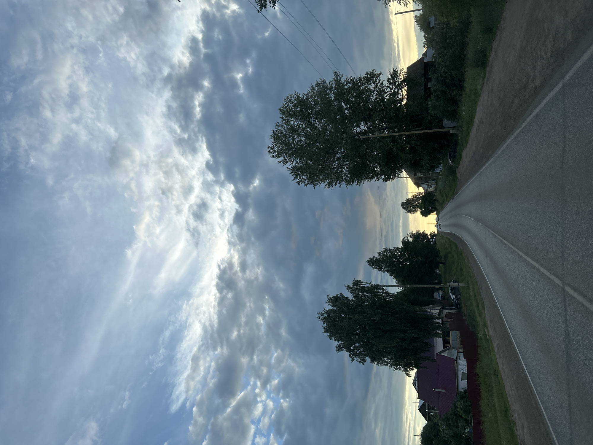The image depicts a straight, paved road winding through a rural area. The road is bordered by grass and trees on both sides, with houses visible in the distance. The sky is filled with dramatic clouds, with a hint of sunset visible on the horizon. The overall mood is peaceful and serene. The image does not provide specific clues about the location within the city of Фоки, Russia, other than it is a rural road. There are no people or specific actions taking place in the scene.
