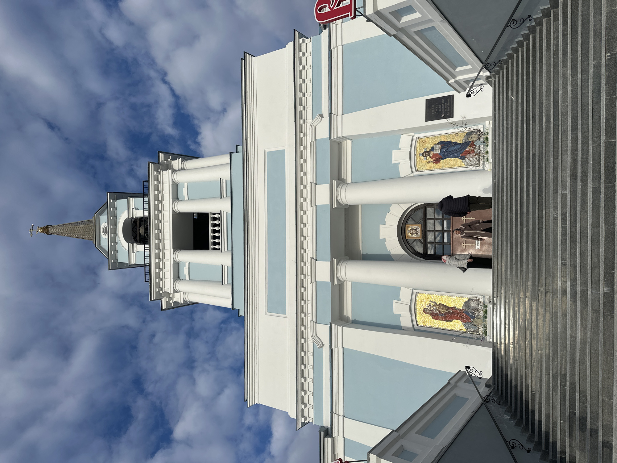 The image shows the Cathedral of the Annunciation in Votkinsk, Russia. The cathedral features a light blue and white exterior with classical columns and mosaic icons adorning its walls. A tall, slender spire topped with a golden cross rises above the main structure. Steps lead up to the entrance. The sky is visible behind the cathedral, showing blue interspersed with white clouds. People are visible near the entrance.