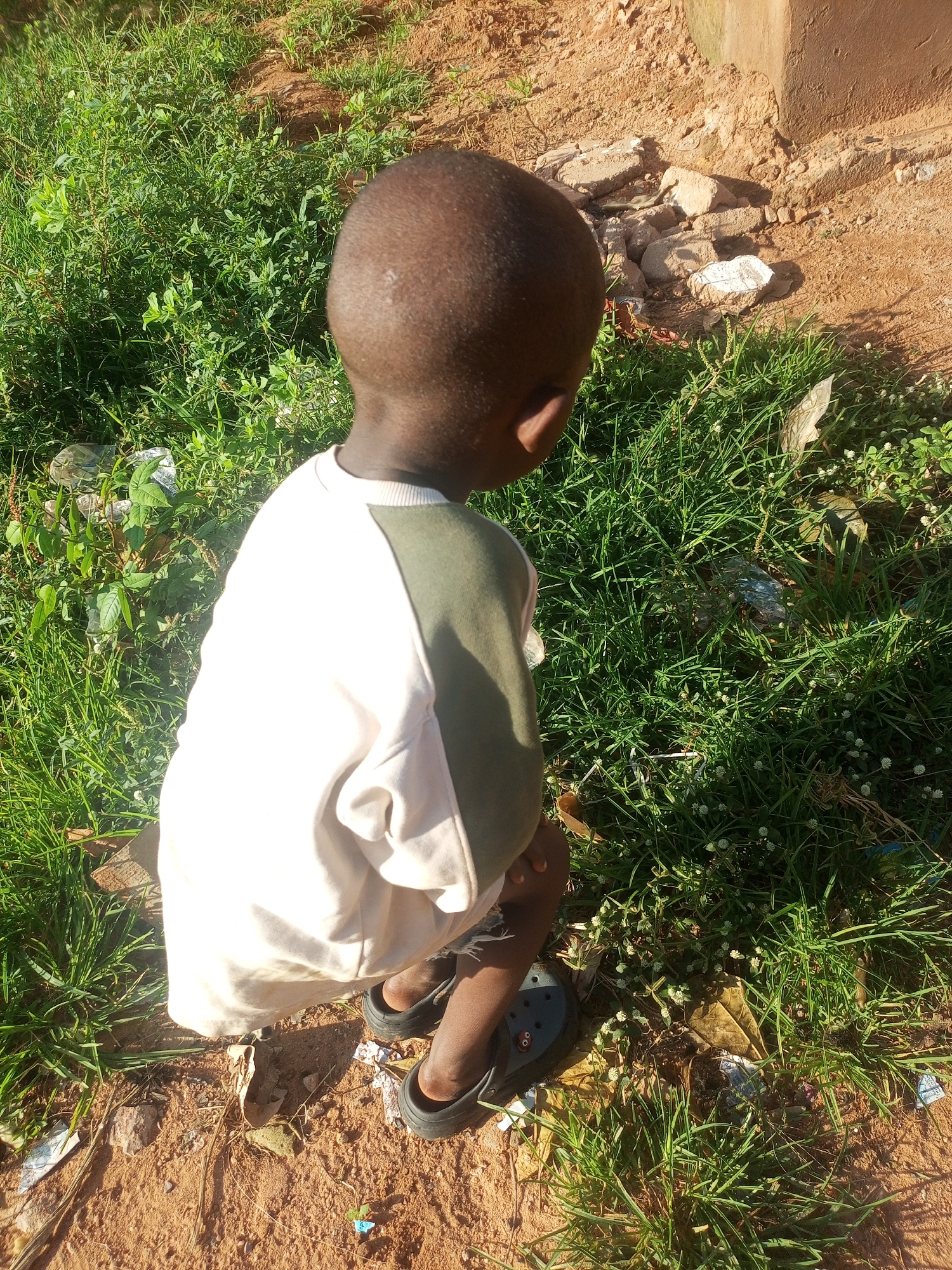 The image shows a young child from the back, wearing a white and green long-sleeved shirt and black crocs, crouching in a patch of tall green grass and dirt. The child's head is tilted downwards, suggesting they are looking at something on the ground. Scattered on the ground are rocks, some leaves, and what appears to be litter, including plastic bags and wrappers. The background consists of more grass and a dirt patch with a concrete structure on the right side, casting a shadow. The lighting indicates it is daytime with bright sunlight. The scene is outdoors in what appears to be a somewhat unkempt area.