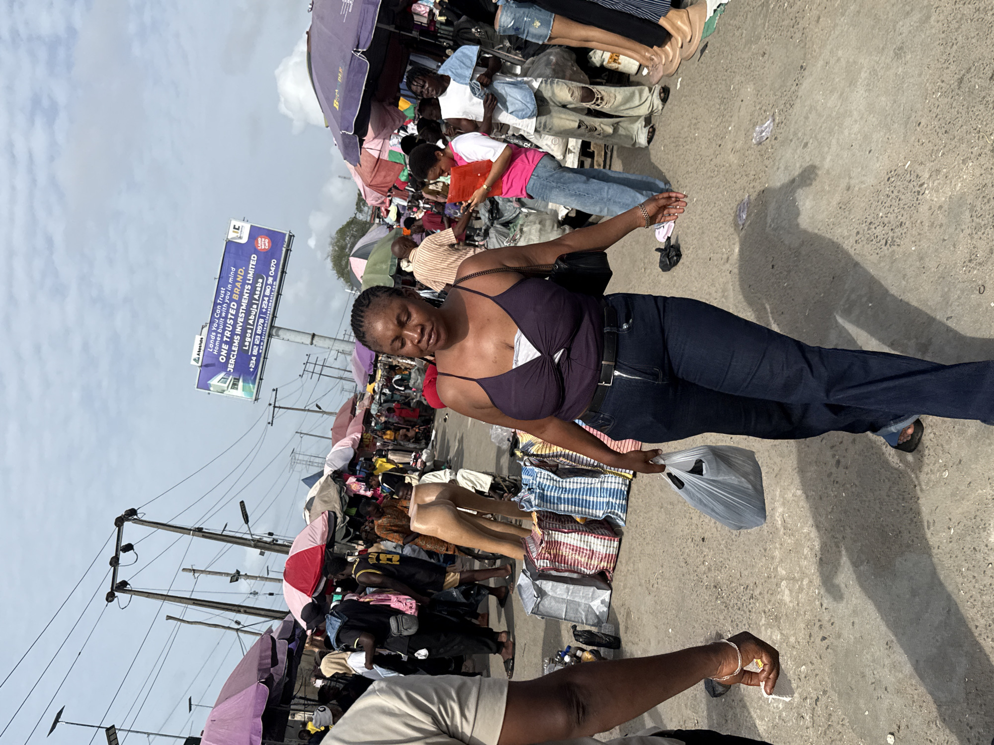 The image captures a street market scene in Lekki, Nigeria. A woman in a purple top and jeans is at the foreground, holding a plastic bag. To her left, there are stalls displaying clothing, and mannequins. Overhead, there are colorful umbrellas providing shade. A billboard advertising Jerclems Investments is visible in the background. Utility poles and wires are visible above the stalls. People walk around the market.
