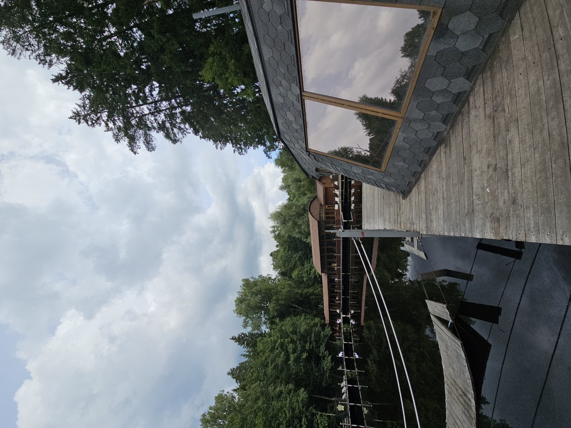 The image depicts a wooden structure with a dark, hexagonal-shingled roof and large, reflective windows. The structure appears to be elevated, as suggested by the wooden walkway leading to it and the view of trees below. A forest surrounds the structure, with dense green foliage and tall trees under a cloudy sky. The reflection in the windows shows trees and clouds, indicating it is daytime.
The structure resembles a modern, possibly vacation-style dwelling or an observation deck. The wooden planks of the walkway and the structure's exterior suggest a natural, rustic setting. The sky is overcast with dramatic, light-colored clouds, implying a partly cloudy or possibly rainy day. The overall mood is serene and natural, with the architecture blending into the forest environment. There is no visible text in the image.
The image is oriented vertically, with the sky occupying the top half and the structure and forest filling the bottom. The water-like surface in the foreground reflects the trees and the structure, adding depth and a sense of stillness to the scene.