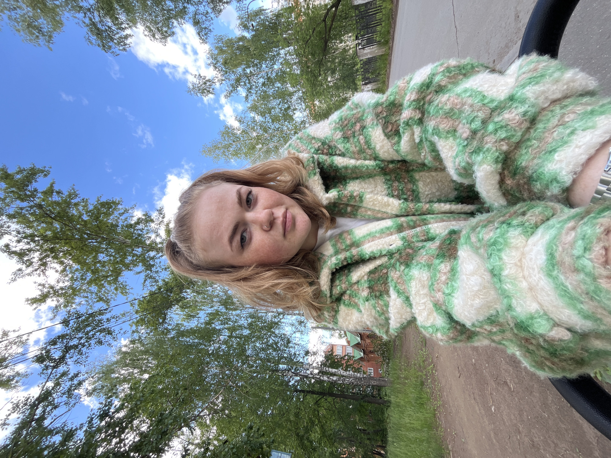 The image is a daytime selfie of a young woman taken outdoors in Chaikovsky, Russia. The woman, who has blonde hair, light skin, and visible freckles, occupies the left side of the frame, looking directly towards the camera with a neutral to slightly smiling expression. A small silver ring is visible in her left nostril. She is wearing a textured, fluffy jacket or cardigan with a striped pattern featuring shades of light green, white, and light brown.
The background behind her reveals a clear blue sky dotted with scattered white clouds, indicating sunny weather. Below the sky, numerous green deciduous trees, some with light-colored trunks suggesting birch, fill the upper and mid-ground, providing a natural setting. A dirt or gravel path is visible along the ground, running parallel to a grey paved surface, suggesting a park, street, or residential area. In the distance, a multi-story building with a reddish-brown roof and light-colored walls can be seen peeking through the foliage. A dark, curved object, possibly a part of a bicycle or stroller handle, is visible in the lower right corner. No text is visible in the image.