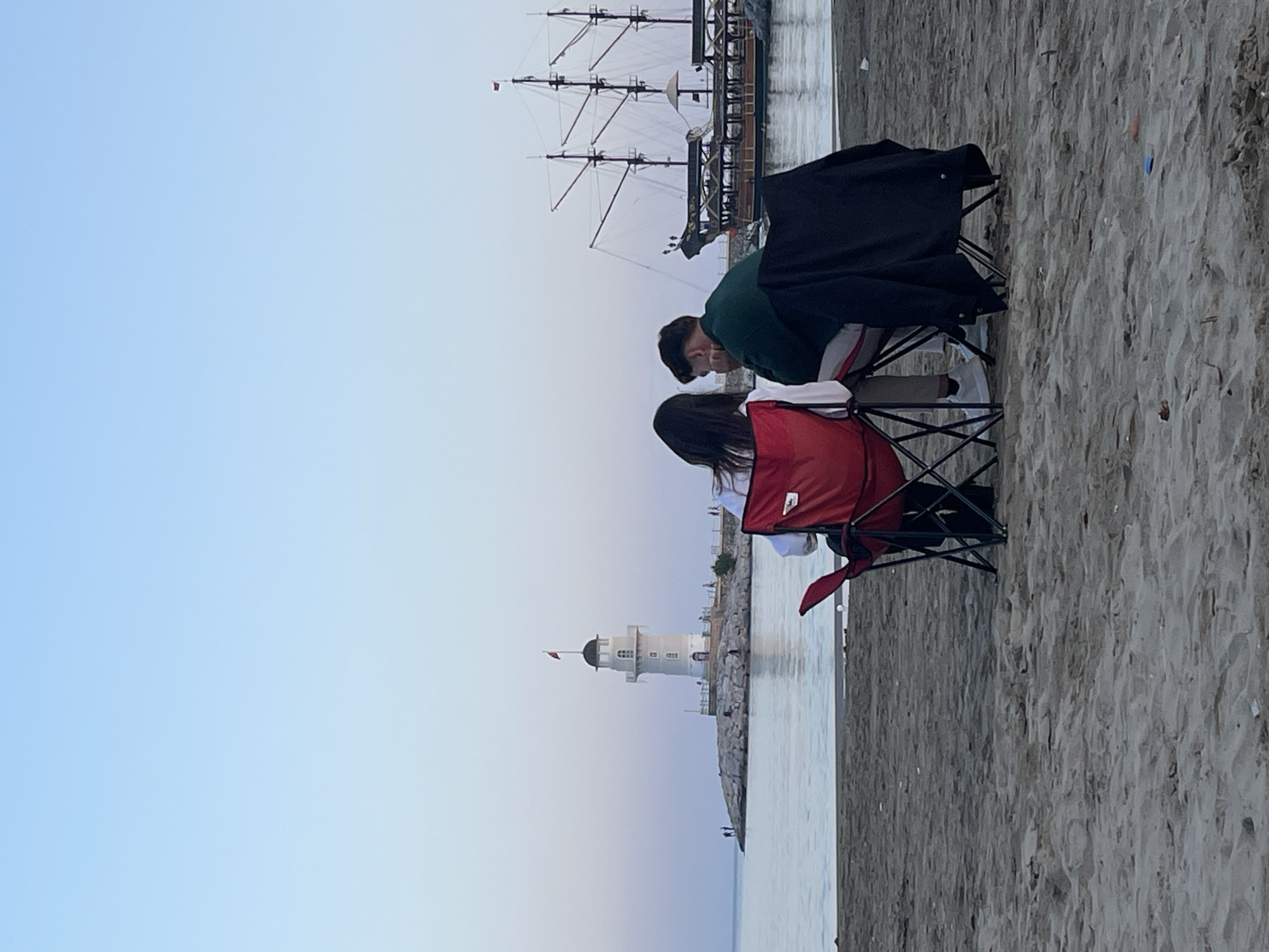 Two people are sitting on portable chairs facing a lighthouse, with a ship in the background. They are located on a beach in Alanya, Turkey. The sky is light blue, transitioning to a light orange near the horizon. The sand is gray.