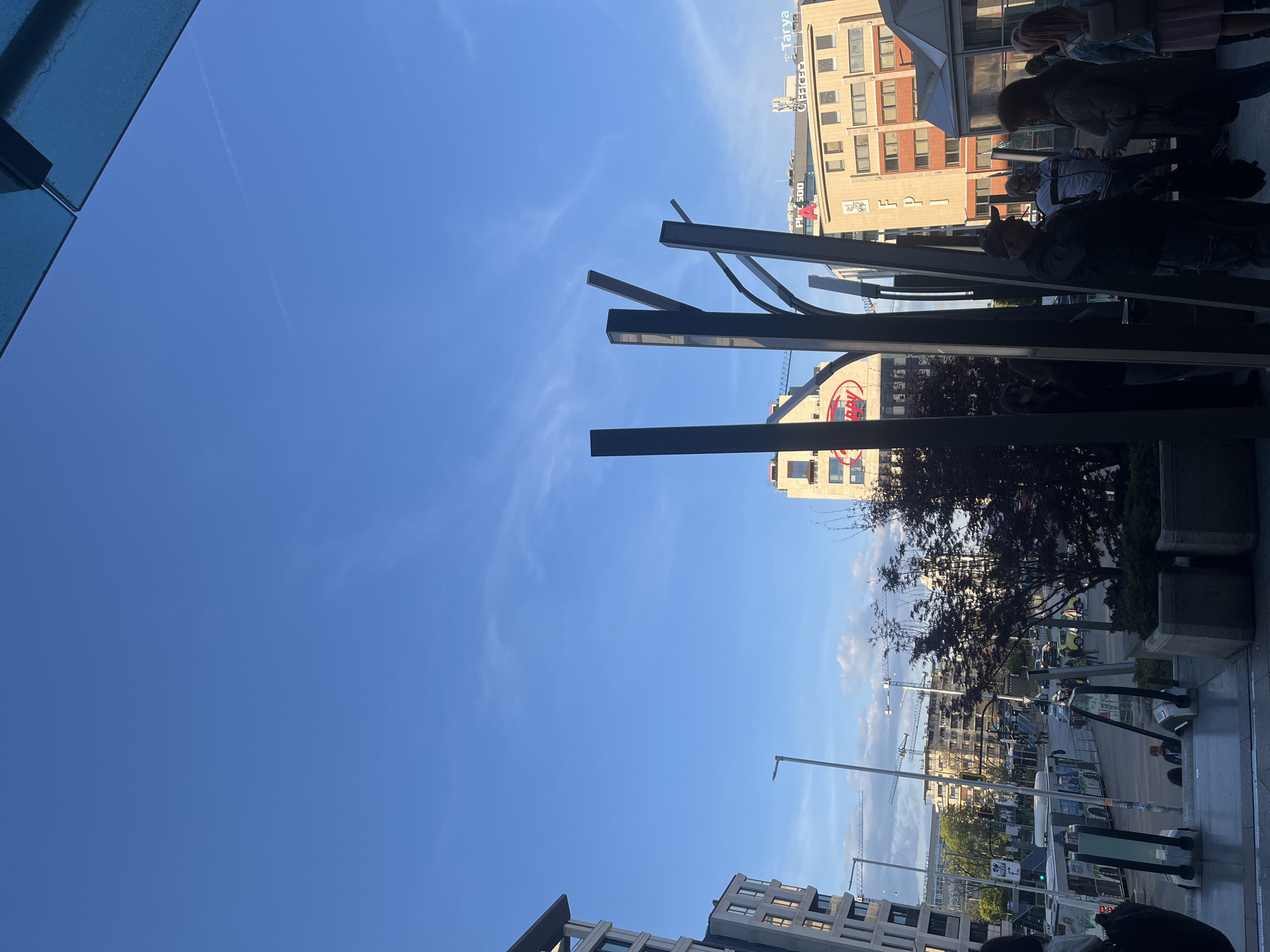 The image shows an outdoor scene in Sofia, Bulgaria. The sky is blue with a few wispy clouds. Various buildings of different heights and architectural styles line the street. Some buildings have signage visible. The image is taken from a slightly elevated perspective, possibly from a cafe or restaurant terrace. People are visible sitting at tables in the foreground. There are decorative metal structures resembling stylized trees. The city street below has a mix of traffic, including cars and buses, and street lights and trees are visible. Construction cranes are also seen in the distance.