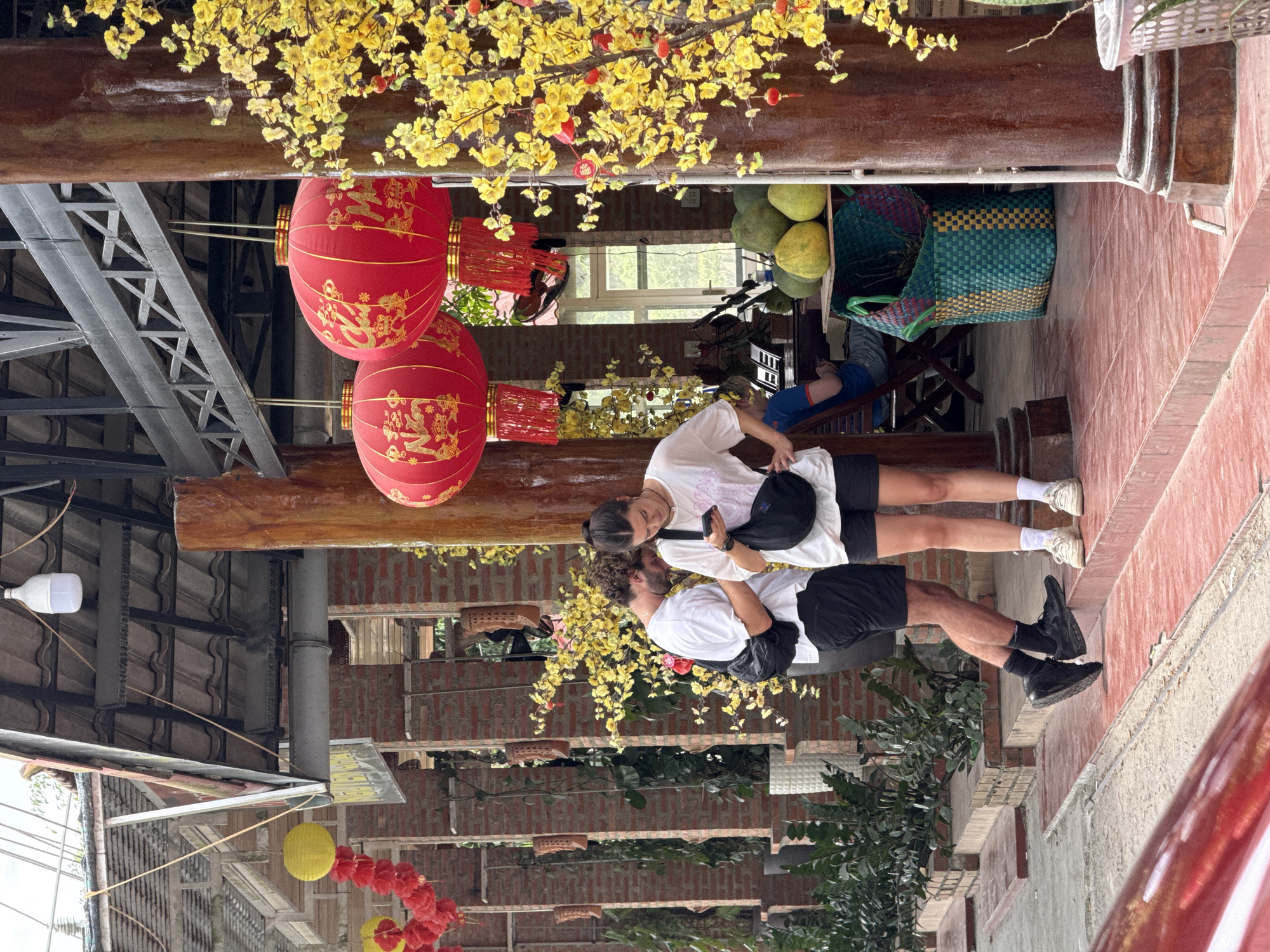 The image depicts a street scene in Nam Khanh Vinh, Vietnam. Two individuals, a male and a female, are standing together near red lanterns and yellow blossoms. Both are dressed in casual attire, with the female holding a phone. The environment consists of buildings with red brick and metallic structures. There are also baskets filled with fruit nearby.