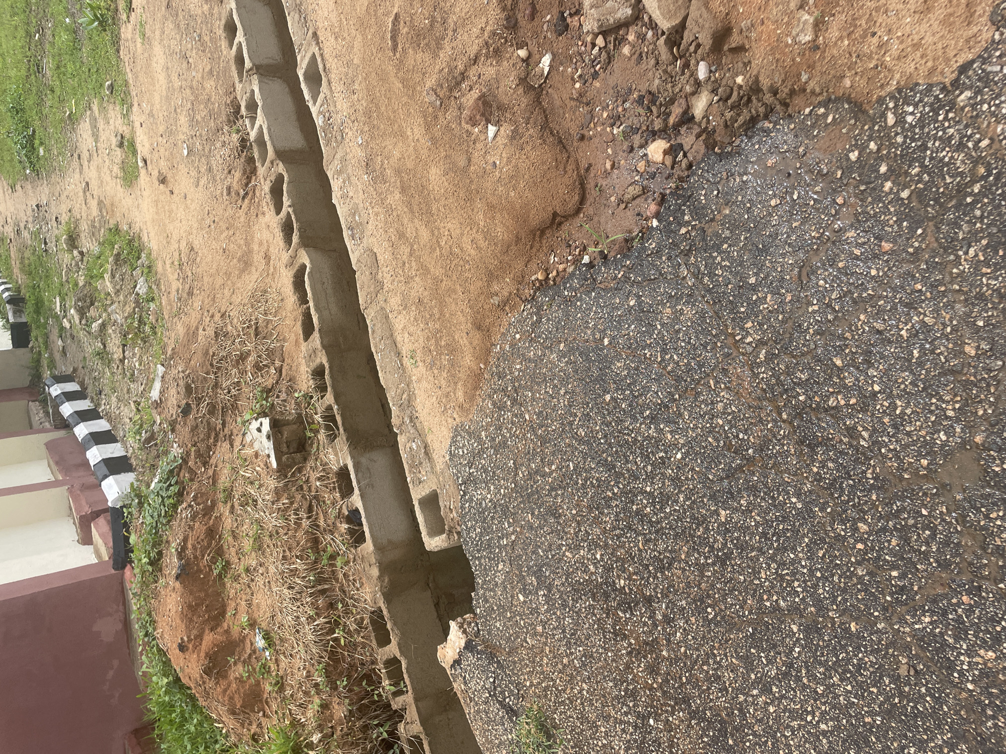 This image depicts an outdoor scene in Lokoja, Nigeria, focusing on a damaged roadway and adjacent construction. The foreground shows a section of dark asphalt road that is broken and appears to be peeling away, revealing exposed aggregate. To the right of the asphalt, a patch of reddish-brown soil is visible, with some small stones and debris scattered on its surface. Further back, a ditch or trench is lined with concrete blocks, suggesting ongoing construction or repair work. The ground is uneven, with patches of dry grass and sparse green vegetation growing in the sandy soil. In the background, a building with a maroon and cream facade is partially visible, with a row of black and white painted concrete blocks lining its base. The overall impression is one of disrepair and ongoing development in an urban or semi-urban environment. There are no people visible in the image.
