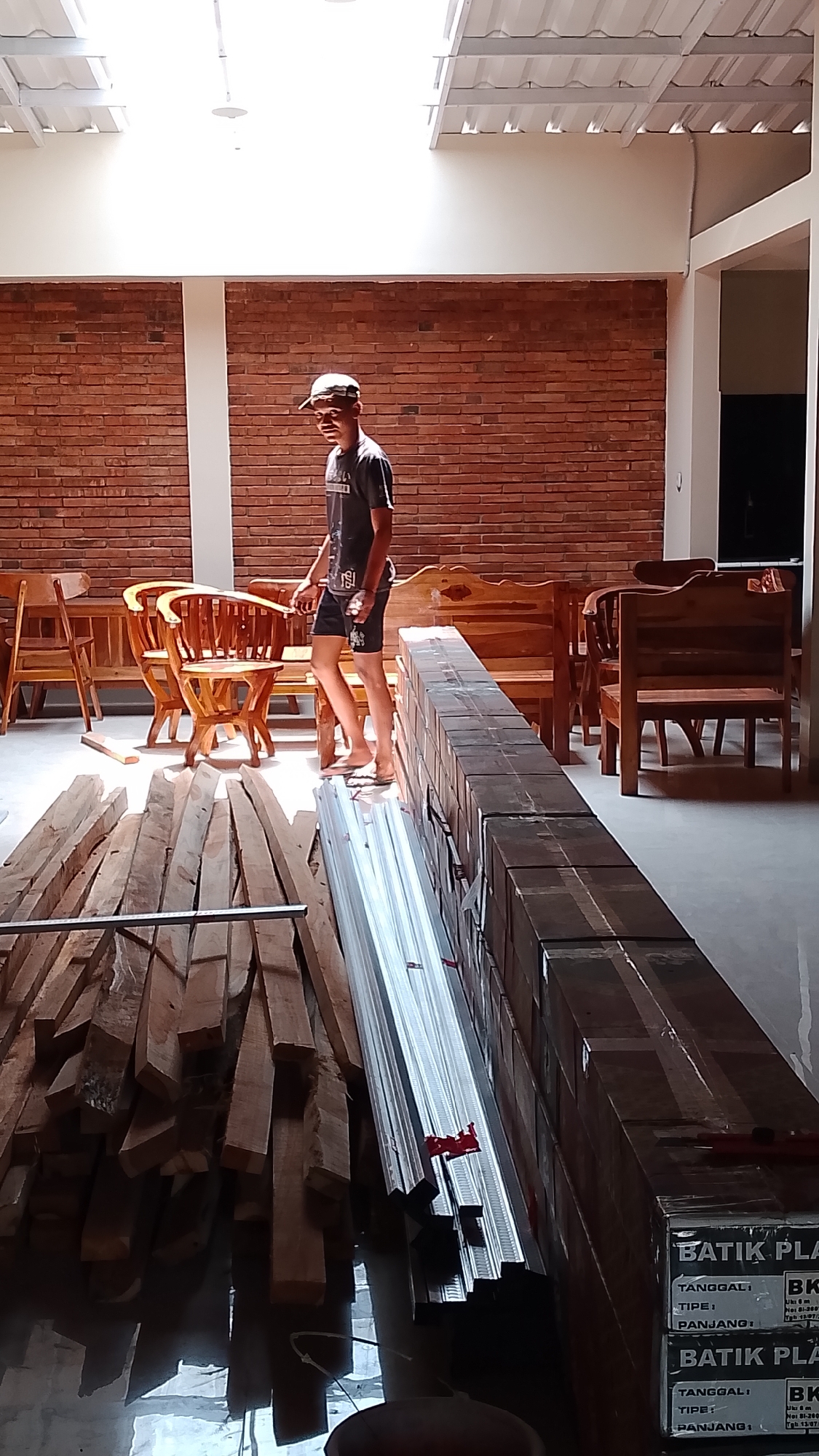 In the city of Karanglangu, Indonesia, a young male wearing a dark t-shirt and a white cap stands near a brick wall. He is wearing dark shorts and flip-flops. The environment appears to be an indoor space with a high ceiling and skylights. In the foreground, there are stacks of wooden planks and long, metallic objects. Behind the person, there are several wooden chairs and benches, suggesting a furniture workshop or showroom. The overall scene depicts a storage or working area for wooden materials and furniture.