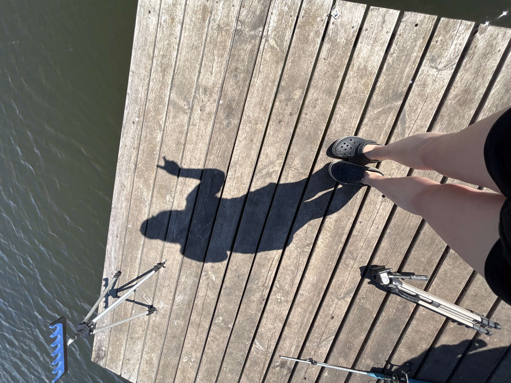 A high-angle, daytime shot shows a person standing on a wooden dock next to water, with their shadow cast on the dock. The person is wearing dark-colored Crocs and dark shorts, with fair skin. A shadow of the person's head and arm is cast on the wooden planks. A fishing rod stand is on the edge of the dock near the water. A tripod is on the dock, near the person. The location is potentially Khryashchevka, Russia, based on the user's specification.