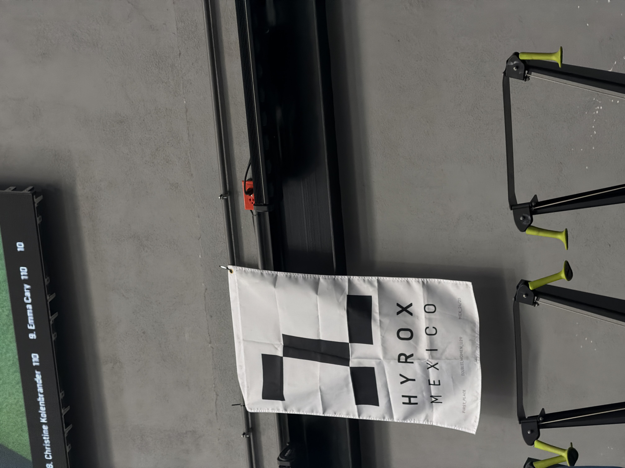 A white flag, featuring the distinctive black "H"-shaped HYROX logo, hangs prominently against a textured gray wall, indicating a fitness competition or event. The flag clearly displays "HYROX MEXICO" in black text, along with "FIRST PLACE," "DOUBLES WOMEN U29," and "MEX 24/25," suggesting a specific category and season for the event.
To the left of the flag, a digital scoreboard is partially visible, displaying competitor information. The visible text includes "8. Christine Kolenbrander 110," "9. Emma Cary 110," and the number "10."
To the right of the flag, parts of several fitness machines, likely rowing machines, are visible. They feature black frames and striking neon yellow handles or footrests. The background consists of a concrete or painted wall, with vertical metal beams and structures. The setting appears to be indoors, under artificial lighting, in San Bernardino Tlaxcalancingo, Mexico.