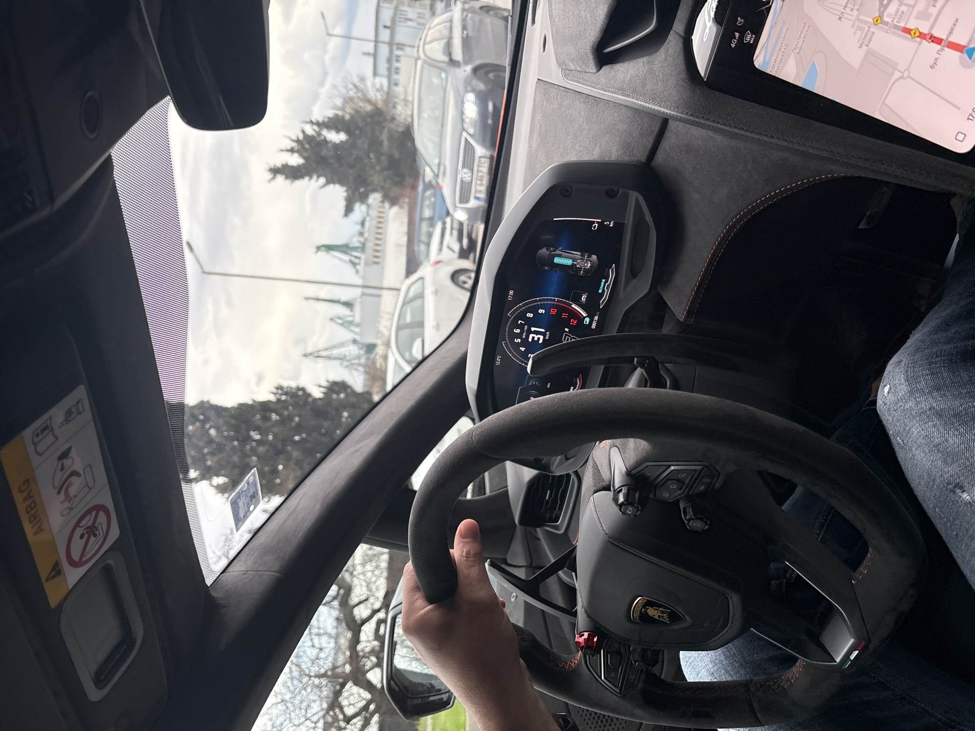 A first-person perspective captures the interior of a high-performance car, likely a Lamborghini Huracán, seen from the driver's seat. The driver's right hand grasps the Alcantara-wrapped steering wheel, which features a Lamborghini emblem, control buttons, and a small Italian flag accent. The digital dashboard display shows an outside temperature of 12.0°C, the time as 17:00 (5 PM), a speed of 31 km/h, and a remarkably low odometer reading of 000136. A battery icon indicates a charge level, with "RECHARGE" also visible, suggesting a hybrid or electric vehicle, or perhaps regeneration.
Through the windshield, an overcast daytime sky is visible. Outside, several cars are parked, one with a Bulgarian license plate "B 6467 TC", confirming the location as Varna. In the distance, large industrial structures, possibly cranes, are prominent. A mobile device, likely a smartphone or tablet, mounted on the dashboard, displays a map application. Visible text on the map includes Bulgarian place names like "Корабостроителна", "Пристанище", "Варна Изток", "Жп лин", "Митница Варна", and "бул. Приморски", all indicative of the port area in Varna, Bulgaria. The passenger-side sun visor features a prominent "AIRBAG" warning label. The driver is wearing blue jeans, partially visible on the right.