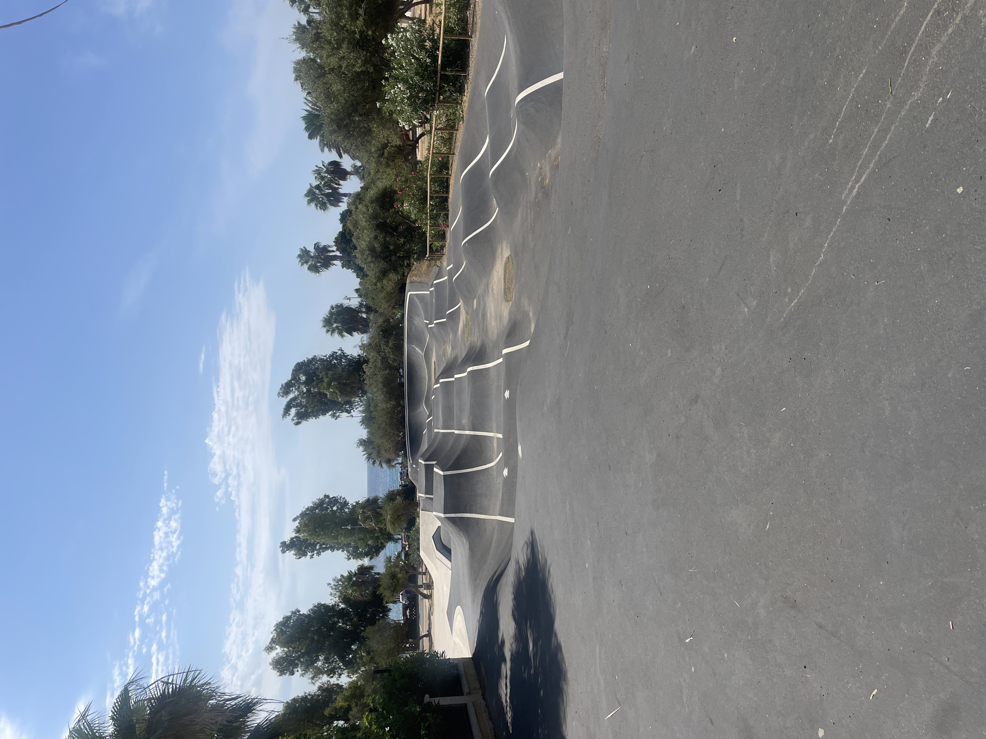 This image shows a public pump track, likely located near the coast in Almuñécar, Spain. The track is made of dark asphalt with white lines marking its features, consisting of a series of undulating mounds and banked turns. In the background, palm trees and other greenery line the area, with a glimpse of the Mediterranean Sea and a beach visible under a clear blue sky with scattered clouds. The foreground is a paved, flat area of asphalt, casting a shadow from an unseen object on the left. There are no people or animals visible in the scene.
