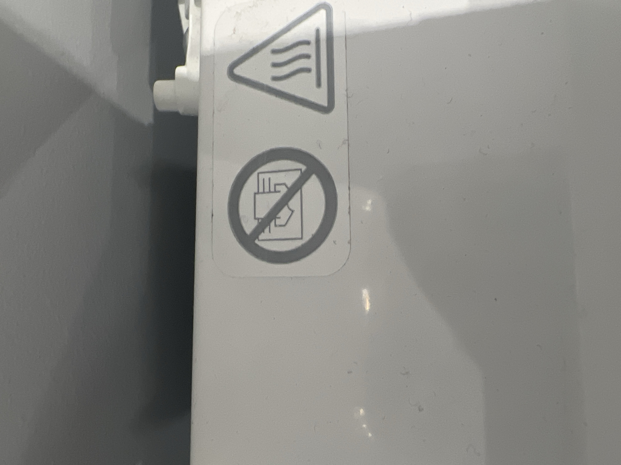 The image shows two safety warning stickers on a white surface, likely a piece of equipment. The sticker on the left is circular with a red border and a diagonal red line crossing a symbol depicting a hand reaching into a device with paper. This symbol indicates "Do not insert hands" or "Do not put hands into the machine." The sticker on the right is a triangle with a black border and a black symbol inside representing three wavy lines above a horizontal line, indicating a "Hot surface" warning. The background appears to be a grey, angled surface, possibly a wall or ceiling. There is a white plastic component visible to the right of the stickers. The scene is indoors, and there are no people or animals present. The overall impression is that of a functional, industrial, or domestic appliance. The location is identified as Podgumer, Bulgaria.