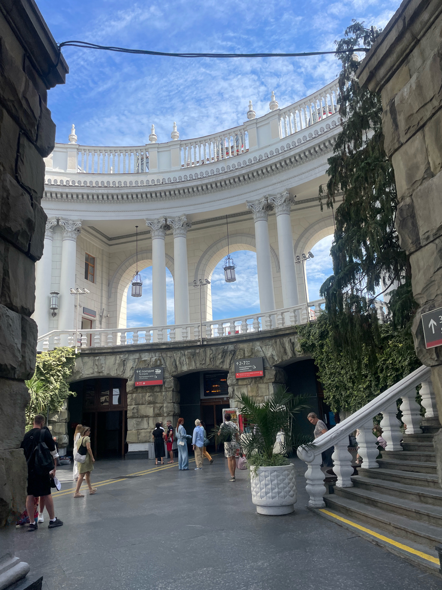 The image depicts the facade of the Sochi Sea Port in Sochi, Russia, a striking example of Stalinist Empire style architecture. The structure features a grand semi-circular building with white columns, arched windows, and ornate balustrades. Several people are visible in the foreground and mid-ground, some walking, others standing, suggesting a public space. The sky above is blue with scattered white clouds. The ground is paved, and there are stone elements and greenery, including a large potted palm, integrated into the scene. Signs with Cyrillic text are present, indicating directions or information relevant to the location. The overall impression is one of architectural grandeur and public activity within a coastal city.